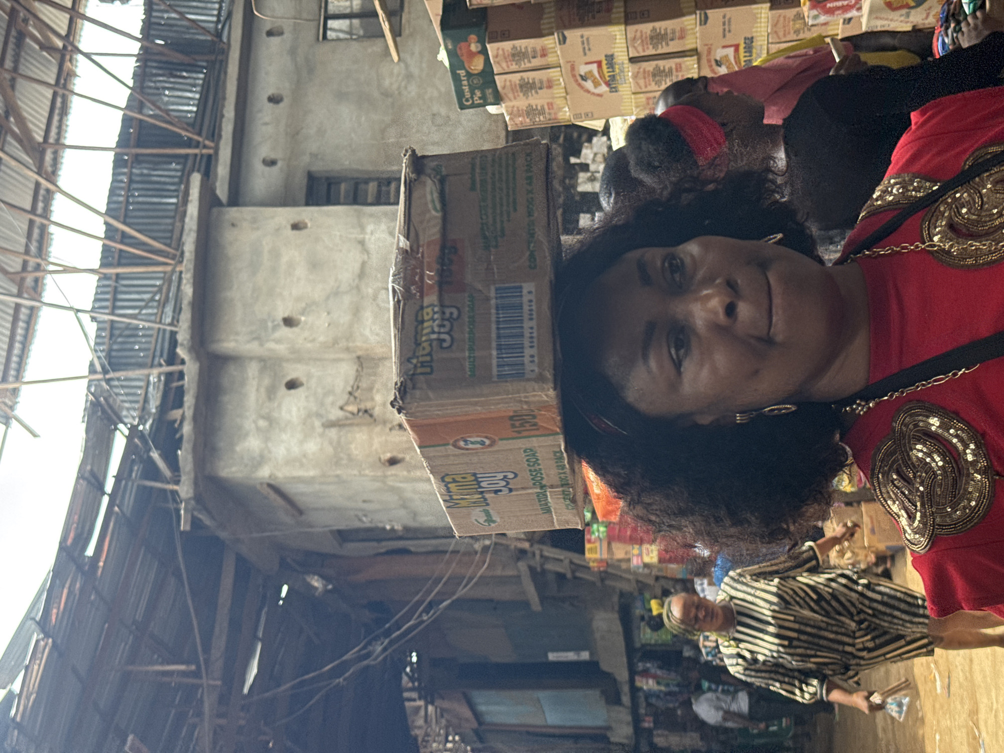 The image depicts a woman wearing a red shirt with gold embellishments, partially obscured by a cardboard box labeled "Ijema Joy MULTIPURPOSE SOAP 1500." Another woman is seen in the background wearing a striped shirt. Various branded boxes such as "Custard Pie", "EXTRA LARGE", and "CABIN" are stacked nearby. The background reveals an urban market scene, possibly in Onitsha, Nigeria, characterized by crowded stalls, overhead roofing structures, and aging buildings.