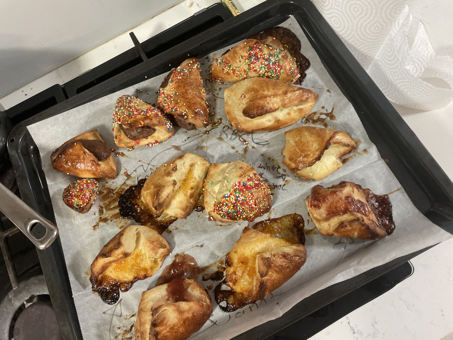 A black baking tray, lined with white parchment paper containing faint handwritten annotations, is presented from an overhead perspective, displaying approximately 16 freshly baked pastries. The pastries are golden-brown and vary in filling and presentation. Several are triangular or crescent-shaped, filled with a dark substance (likely chocolate or a similar spread) and generously adorned with multi-colored sprinkles. Other pastries contain an amber-colored, jam-like filling that has partially oozed and caramelized onto the parchment paper. The tray is situated within what appears to be a home kitchen environment in Raanana, Israel. A portion of a black stove cooktop with metallic grates and a metal pot handle is visible to the upper left. To the right, a white countertop or appliance surface displays a partially visible roll of white paper towels and a small yellow-labeled object. No individuals are present. The pastries are consistent with items commonly prepared or purchased from bakeries in Israel.