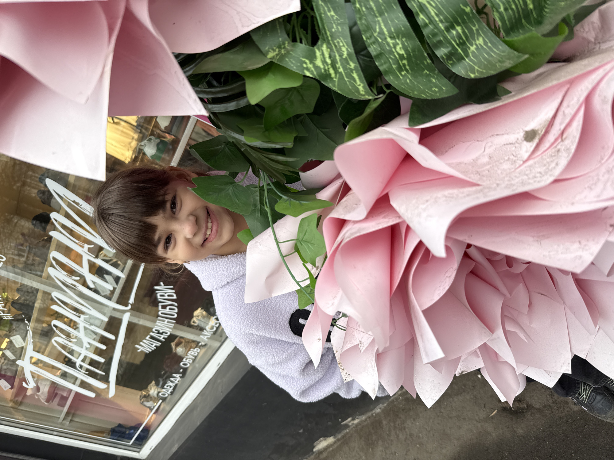 A young girl with brown hair and bangs smiles from behind a large bouquet of pink paper flowers and green artificial foliage. She wears a light purple hooded jacket with a small black and white animal design on the chest. The bouquet obscures much of her body. The scene appears to be outdoors, in front of a store window with white lettering in Cyrillic, reading "МАГАЗИН ОБУВИ - ОДЕЖДА - ОБУВЬ - АКСЕ". The location is Zhukovo, Russia. The girl stands on a wet, gray pavement. The bouquet is so large that it extends beyond her and is partially touching the ground. A pair of black shoes with light-colored laces is visible near the bottom right corner of the frame.