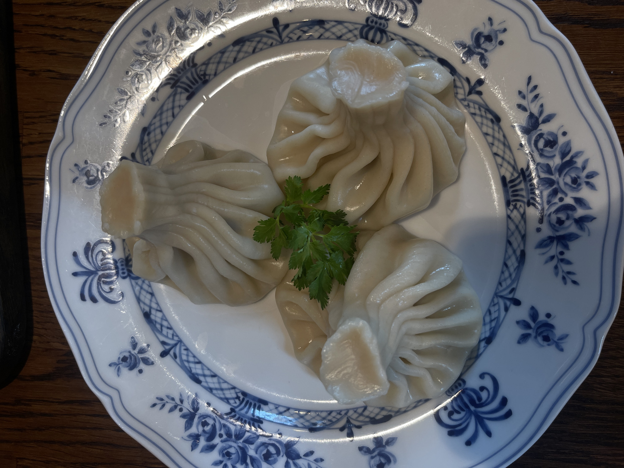 The image shows a plate of three dumplings, likely Khinkali, a type of Georgian dumpling, arranged on a white plate with a blue floral pattern. A small sprig of green herbs, likely parsley or cilantro, is placed in the center, between the dumplings. The plate rests on a wooden surface. There is no visible evidence of the location being Izhevsk, Russia.