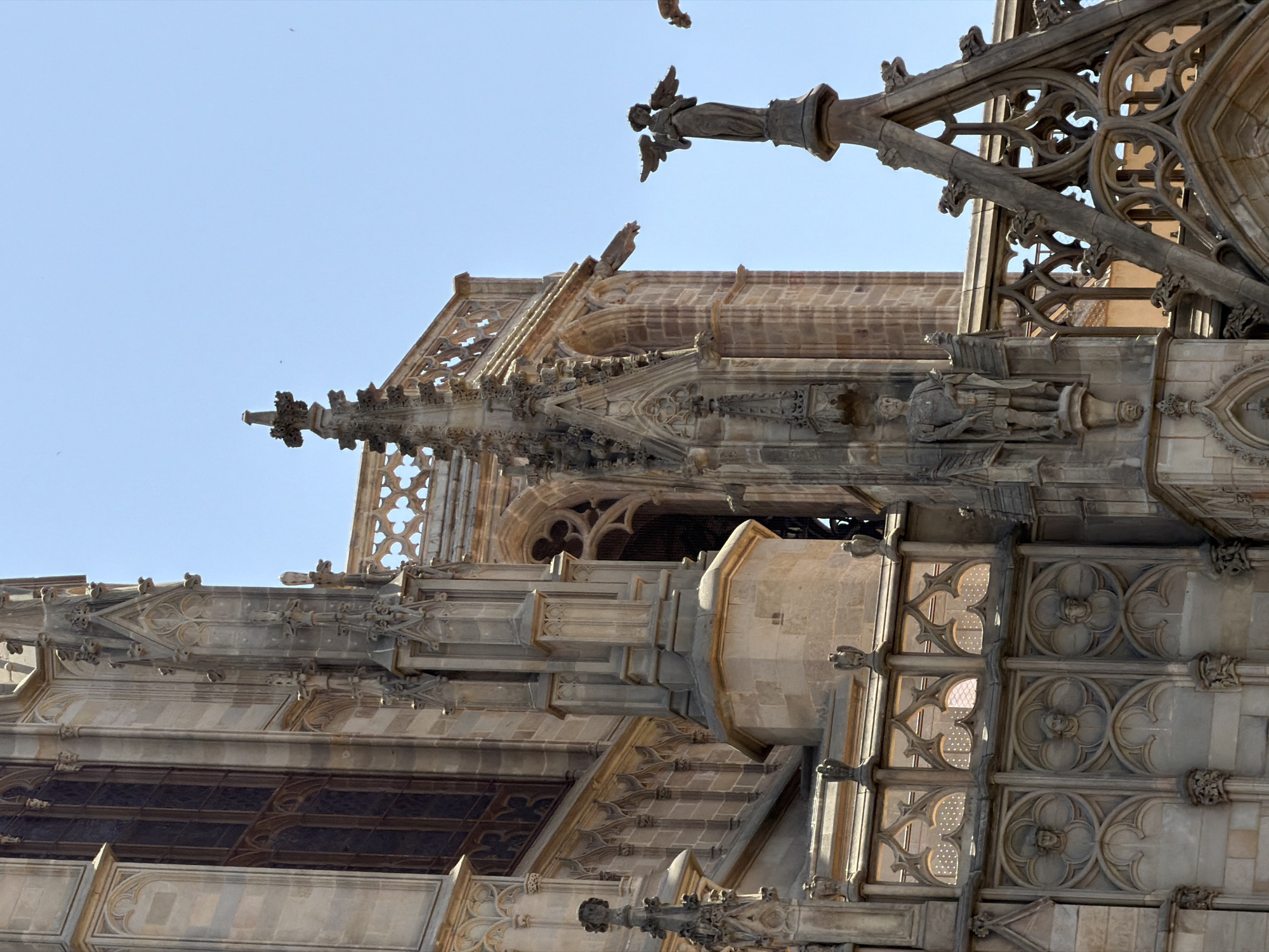 The image shows a close-up view of the architectural details of the Barcelona Cathedral, located in the Gothic Quarter of Barcelona, Spain. The scene focuses on the upper portions of the cathedral's exterior, highlighting intricate stonework, sculptures, and decorative elements against a clear blue sky.
On the right side of the image, a weathered statue of an angel with outstretched wings stands atop a pointed spire. The angel is depicted in a classical style, with flowing robes and a serene expression. Below the angel, gothic spires and tracery are visible, showcasing the detailed craftsmanship of the cathedral. Gargoyles, characteristic of gothic architecture, protrude from the building's ledges, serving as decorative waterspouts.
To the left, a sculpted figure of a man in armor, likely a saint or a historical figure, is mounted on a pedestal. This figure also exhibits classical influences in its design. Further left, a large, tall window with dark panes is visible, framed by ornate stone carvings. The lower part of the cathedral features a series of decorative panels with circular medallions containing sculpted heads, set within pointed arches.
The overall impression is one of grandeur and historical significance, with the sunlight casting sharp shadows and emphasizing the textures and details of the ancient stonework. The clear sky suggests a bright, sunny day.