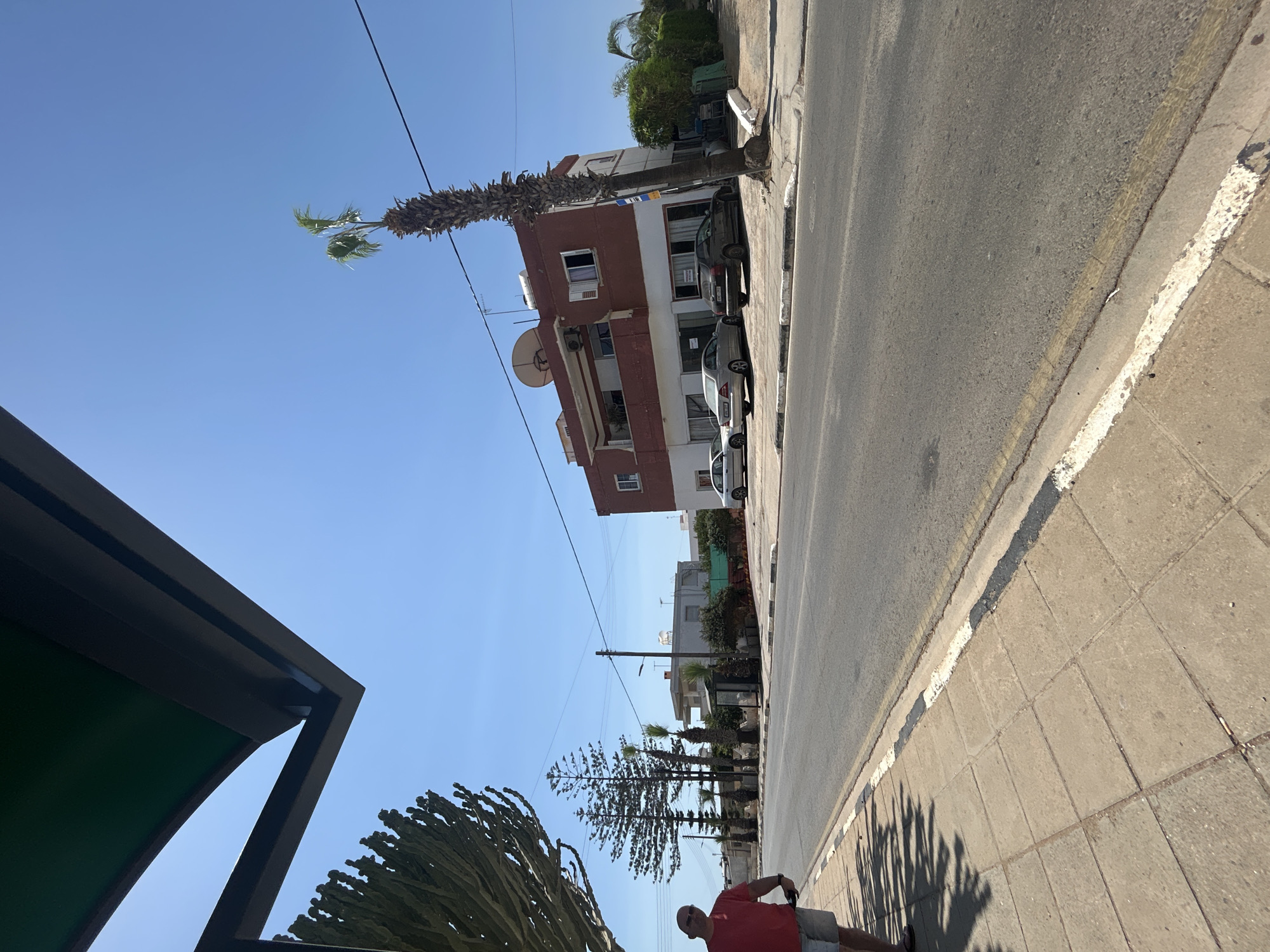 The image shows a sunny street scene in Xylofagou, Cyprus. A man wearing a red t-shirt and grey shorts is walking on the sidewalk on the left side of the image, casting a shadow from overhead foliage. In the background, across the road, a multi-story building with a reddish-brown facade stands. Several cars are parked in front of this building. A tall palm tree is situated in front of the building, and other trees and greenery are visible. The street is paved, and a sidewalk runs along the side. Power lines are strung across the clear blue sky. The overall environment suggests a typical day in a Mediterranean town.
