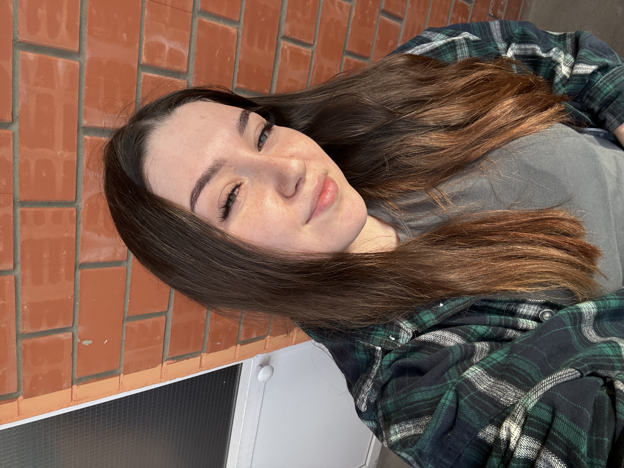 A young woman with long, dark brown hair and light green eyes is taking a selfie outdoors. She is wearing a grey t-shirt and an open green, black, and white plaid flannel shirt. Her skin has a few freckles. She is standing in front of a red brick wall, with a white door and part of a window visible to her left. The lighting suggests it is daytime. The image does not contain any specific information that would allow for identification of the location within the city of Perm, Russia.