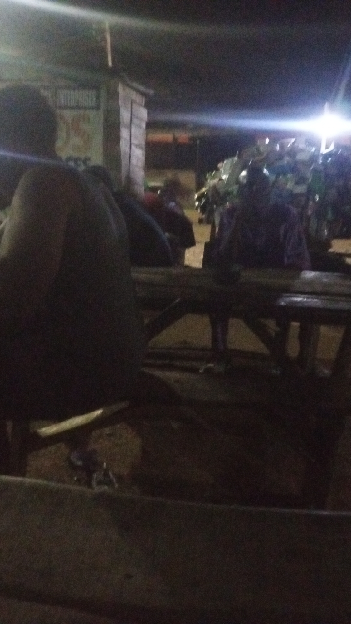 The low-light image captures a night scene in what appears to be a casual outdoor or semi-outdoor gathering area in Adankolo, Nigeria. In the foreground, the back of a person with dark hair, wearing a dark, sleeveless garment, is visible, seated at a rustic wooden table with a corresponding bench.
Further back, other individuals are partially seen, also seated around similar wooden tables, engaged in what seems to be socializing or waiting. To the left, a simple wooden structure, possibly a kiosk or small building, is present. A sign on this structure clearly displays the word "ENTERPRISES" at the top, with fragments of other words like "DS" and "CES" visible below it.
The background is dominated by intense, artificial bright lights that cause lens flare, illuminating a crowd of people and several vehicles, possibly motorcycles or tricycles, indicating a busy public space. The overall impression is that of a local evening hangout or market area.