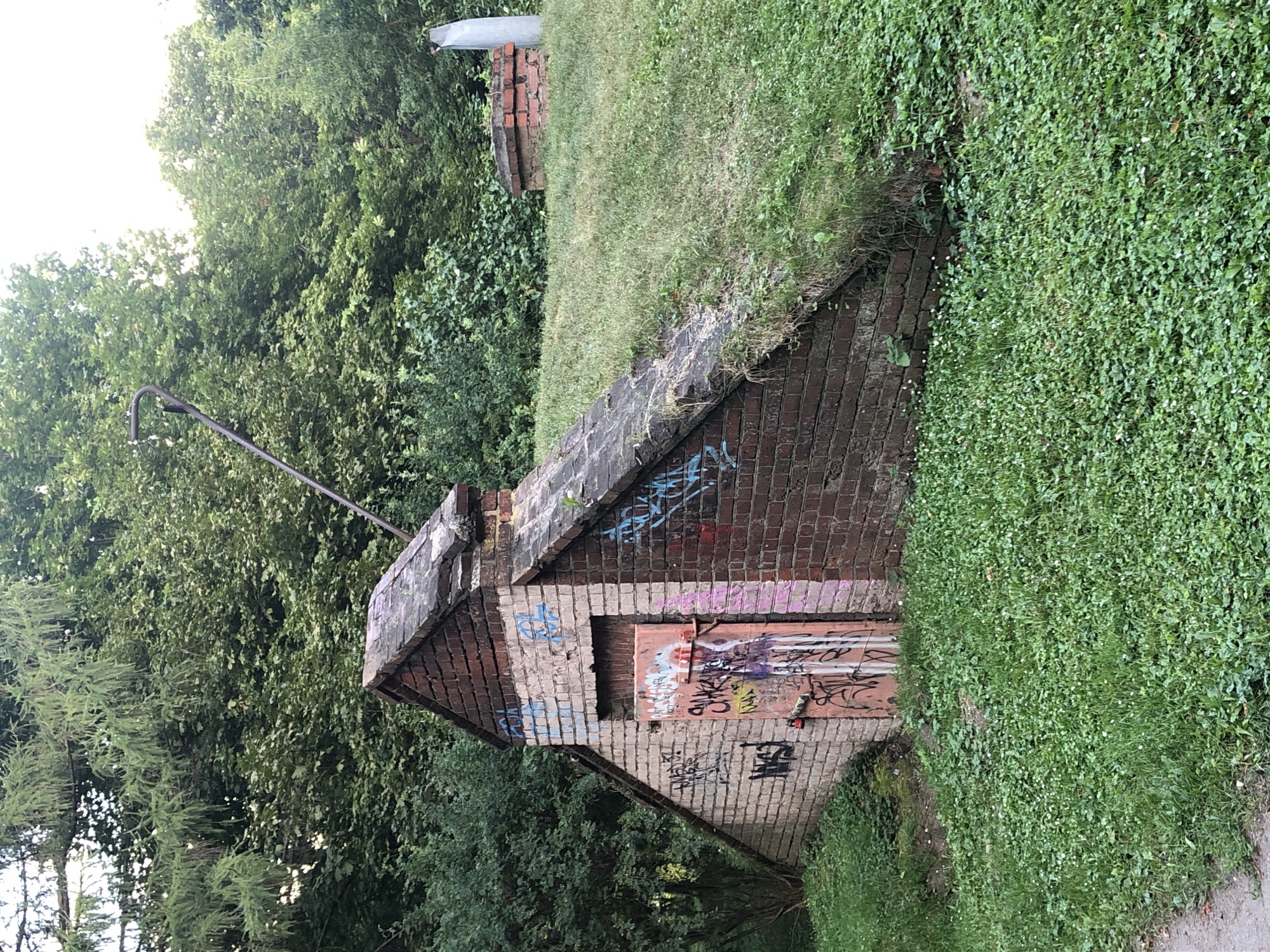 An image shows a partially underground brick structure covered in grass on a sunny day. The structure is a small, triangular building with a sloped roof and a red-painted door covered in graffiti. The building is situated on a grassy hill surrounded by trees. In the background, a brick chimney and a silver metal object are visible. The environment suggests an outdoor, possibly park-like setting.