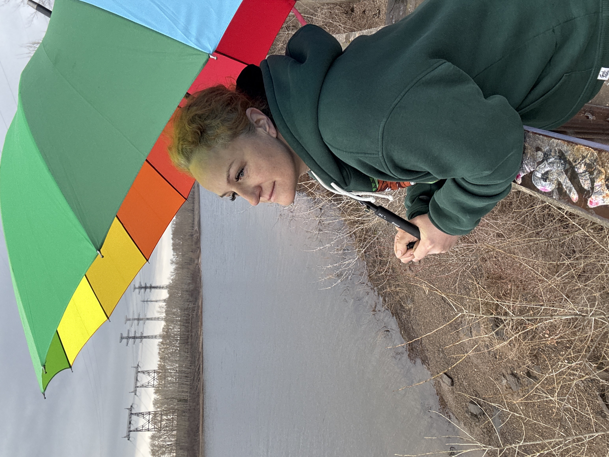 A woman in a green hooded sweatshirt stands near a body of water holding a rainbow-colored umbrella. In the background, several electrical towers are visible along the distant shoreline. The environment appears to be a riverbank with sparse vegetation and overcast skies. The location is likely the city of Chaikovsky, Russia, based on the prompt. The woman is looking down. She is holding the umbrella in her right hand. A metal railing is present.