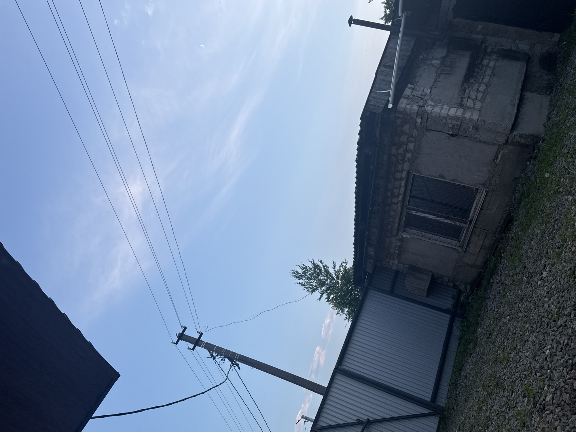 The image depicts an outdoor scene with a clear sky. In the foreground, a grey corrugated metal fence runs horizontally across the left side of the frame. To the right of the fence, a dilapidated, single-story structure is visible. This building appears to be constructed of concrete blocks and has a dark-colored, corrugated roof. A window with metal bars is present on the facade of the building. The ground in front of the fence and building is covered with gravel and sparse vegetation. A utility pole with multiple wires extends upwards into the sky from behind the fence. In the background, a deciduous tree stands tall. The overall environment suggests an industrial or semi-urban setting, possibly a yard or storage area in Birobidzhan, Russia.