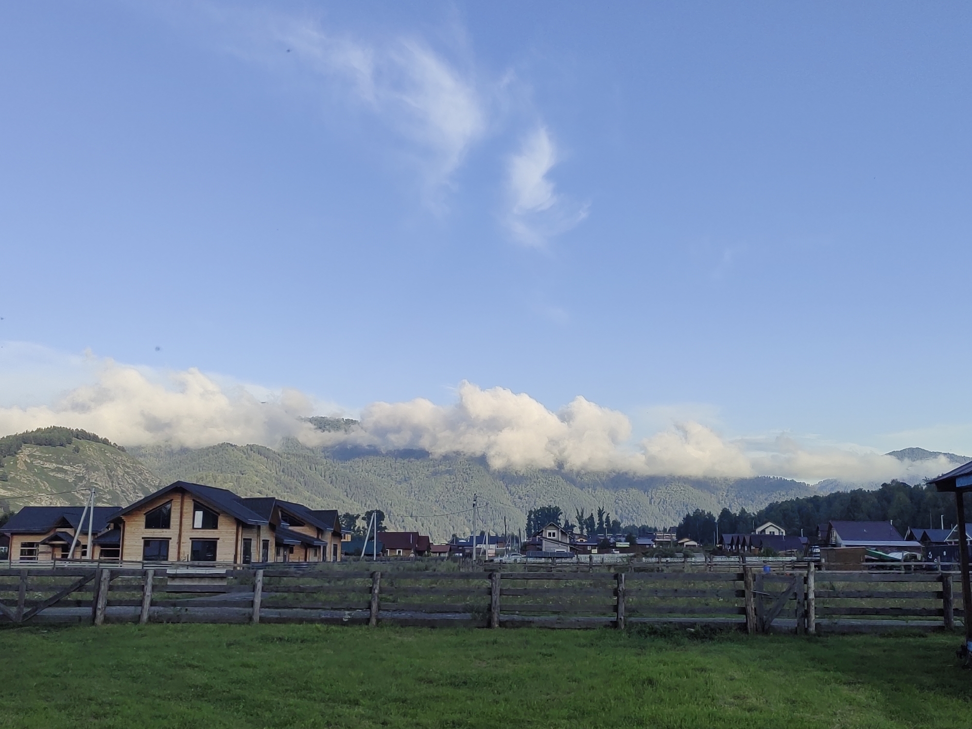 A wide, clear shot shows a village with mountains behind it under a bright blue sky with scattered white clouds. The foreground is a vibrant green lawn with a rustic wooden fence. Behind the fence, a few houses with wooden exteriors and dark roofs are visible, along with other buildings and structures indicating a rural settlement. The mountains in the background are covered in lush green trees, and some clouds hug their peaks. The scene appears to be in Anos, Russia, based on the context provided.