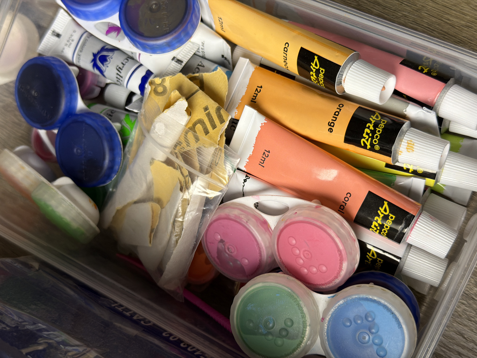 A close-up, high-angle shot reveals a collection of art supplies stored within a clear plastic container, likely situated in Zvezdara Urban Municipality, Serbia. The container is filled with numerous tubes of paint, primarily acrylics, displaying a vibrant palette of colors including yellow, orange, coral, pink, blue, green, and white. Many tubes are branded "pepco® Artist" and indicate a volume of "12ml." Some tubes are open, showing paint at the nozzle, while others appear unused.
Alongside the paint tubes are several small, round pots of paint, possibly watercolors or tempera, with clear lids revealing bright pink, green, and blue pigments. One green lid partially displays the text "USE AFTER EACH USE." Scattered among these items is some crumpled yellow and white paper or packaging material, possibly from art supplies, featuring partial words like "min" and "8." Blue plastic caps, distinct from the paint tubes, are also visible.
No specific activity is depicted, but the arrangement suggests a collection prepared for or stored after artistic endeavors. The items are neatly contained, implying organization within a creative space. Visible text on the tubes and packaging includes "pepco® Artist," "12ml," color names such as "carneos," "orange," and "coral," along with fragments like "Zoll SNAC," "crylic," "My color," "you," "paus.oz," and "140002 CATTI." The scene is brightly lit, likely indoors, with a subtle wooden texture visible in the background.
