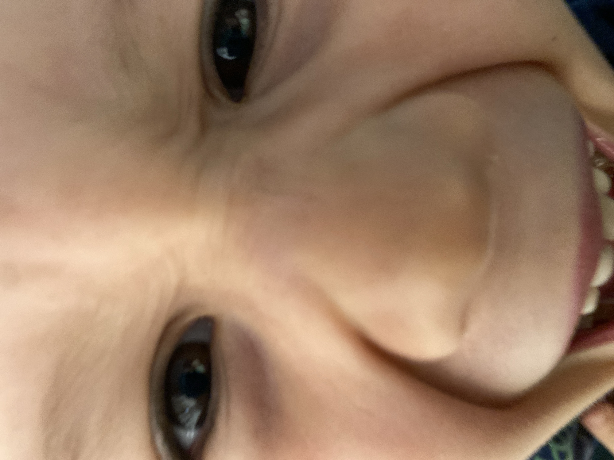 A close-up, slightly rotated image shows a child's face. Two dark eyes are visible, with light reflecting in them. The skin is fair, with creases around the eyes and cheeks. The child's mouth is slightly open, revealing some teeth and a portion of the tongue. The background is blurry and indistinct, with a hint of blue and green patterns. There is no indication that this photo was taken in Chaikovsky, Russia.