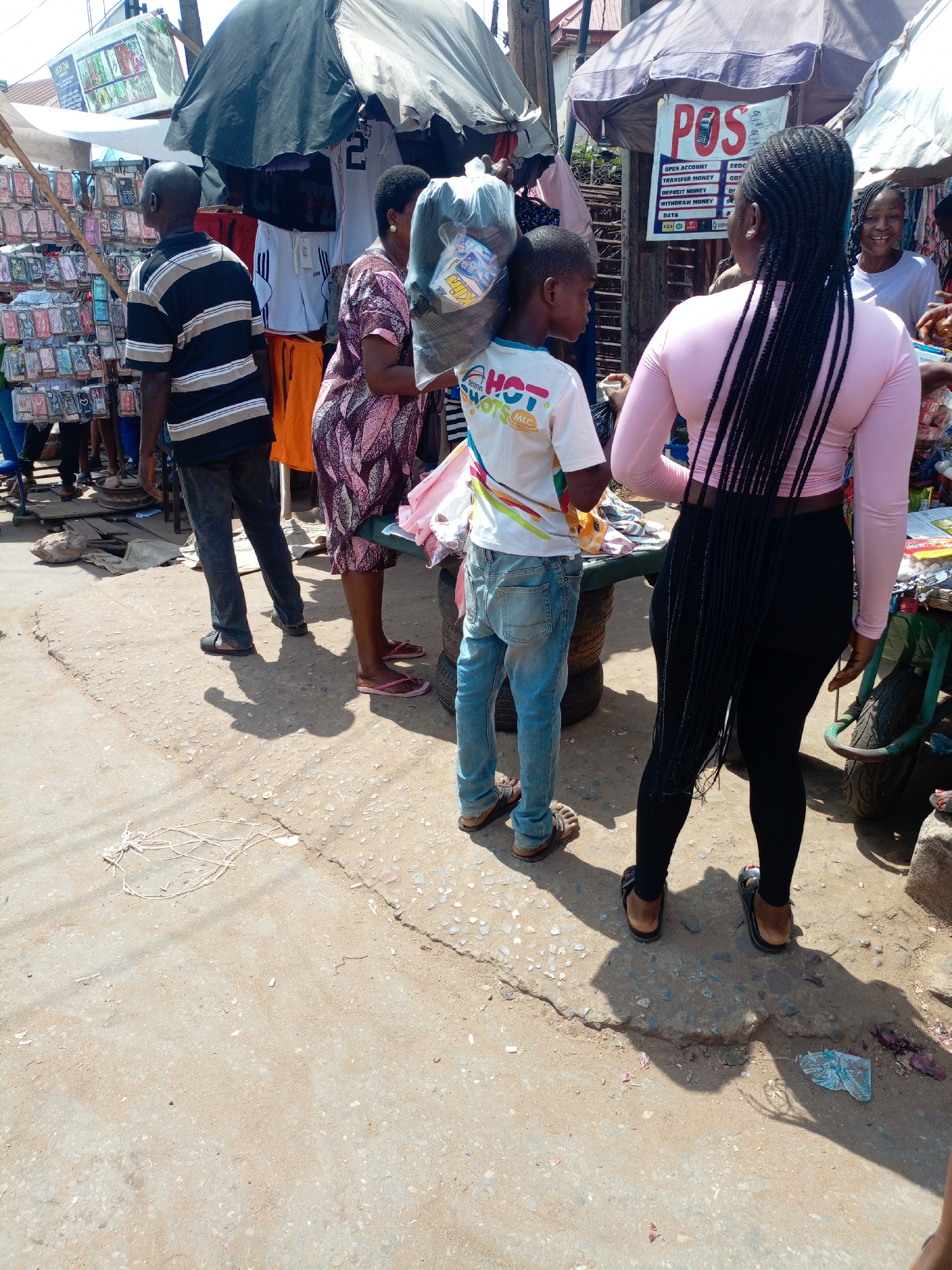 This daylight image captures a vibrant outdoor market scene in Onitsha, Nigeria, under bright sunlight that casts distinct shadows on the ground. Several individuals are present, engaging in various market activities.
On the left, a man in a black and white striped shirt and dark pants stands with his back to the camera, looking at a stall packed with numerous phone cases. Next to him, a woman in a patterned dress is partially visible, also facing a stall. A young boy, wearing a white t-shirt with "HOT SHOTS MLC" text and blue jeans, holds a large transparent plastic bag over his shoulder.
Further right, a woman with long black braided hair, dressed in a pink long-sleeve top and black leggings, stands with her back to the camera. She appears to be at a Point of Sale (POS) service stall. Behind her, another woman with a smiling expression is partially visible. The market stalls are shaded by large, dark umbrellas. One stall displays clothing items, including a white jersey with the number "2" and orange shorts.
A prominent banner for the "POS" service lists various transactions: "OPEN ACCOUNT," "TRANSFER MONEY," "EEDC," "GOY," "DEPOSIT MONEY," "WITHDRAW MONEY," and "DATA." The ground consists of cracked concrete and dirt, with some litter such as plastic and string. The scene conveys a bustling, functional market environment.