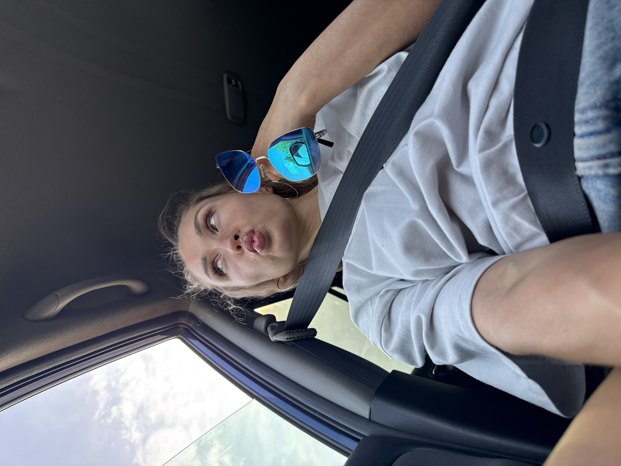 A young woman with fair skin is in a car wearing a white t-shirt and a seatbelt. She is holding blue-tinted sunglasses. Her lips are puckered. The car's interior is blue and black. The location is possibly Chaikovsky, Russia, but there is no confirmatory evidence within the image.