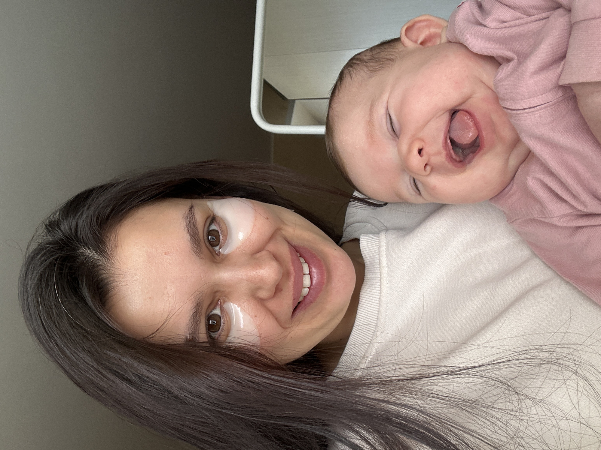 A woman with dark hair and eye masks smiles at the camera. A baby in pink clothing, eyes closed, mouth open, rests on her shoulder. The background is blurred, showing a wall and a white structure. It's impossible to confirm the location in Chaikovsky, Russia based on the visual information.