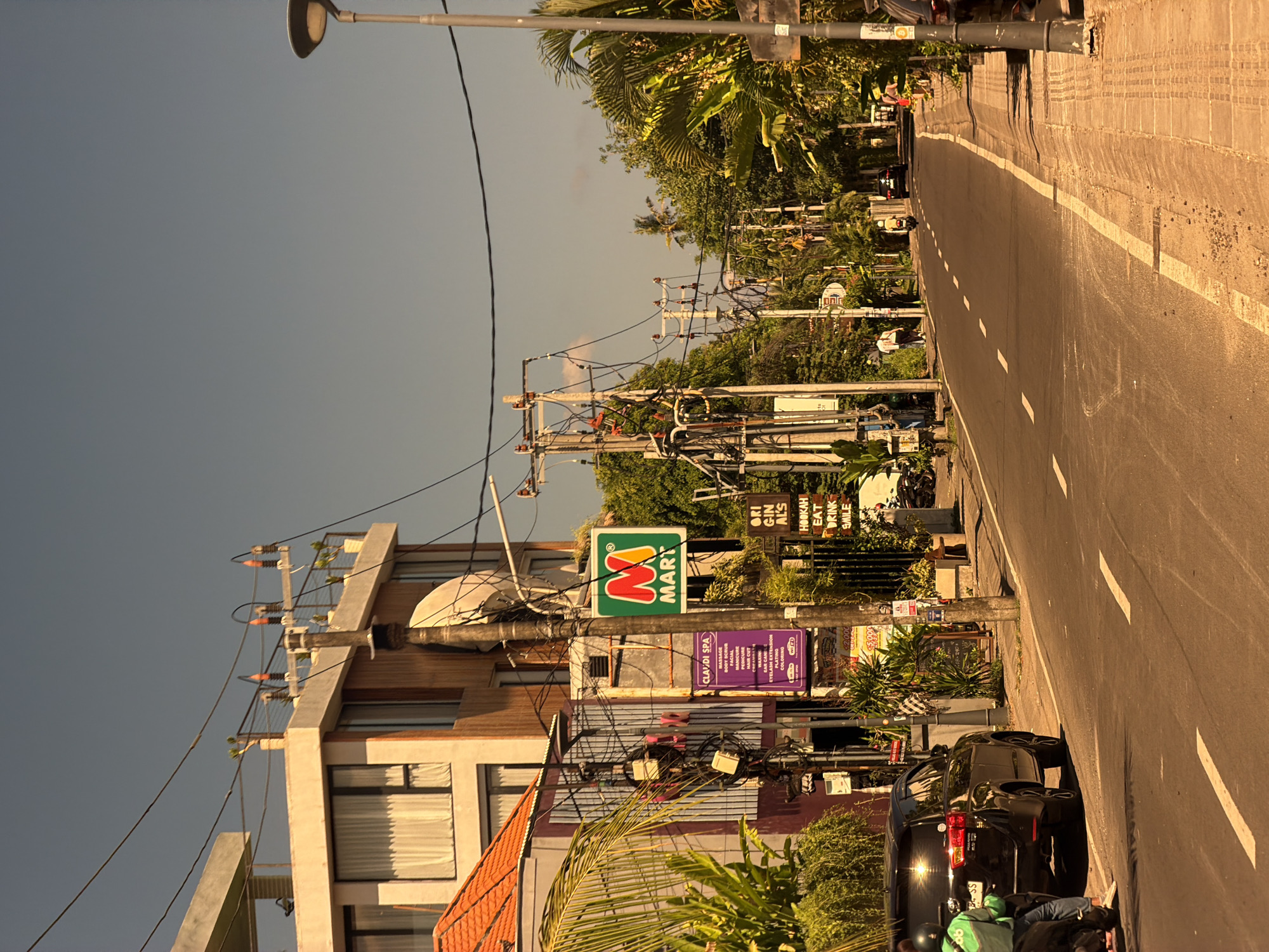 The image shows a street scene in North Kuta, Indonesia. The photo is taken from a sideways perspective, showing a road with a white dividing line. On the right side of the road, there is a parked black SUV. A person wearing a green shirt and dark pants sits on the pavement next to the car. The left side of the street has several buildings with storefronts, including an "M Mart" convenience store, a spa called "CLAUDI SPA", and a sign that reads "ORI GIN ALS" and "HOOKAH EAT DRINK SMILE". Overhead, there are numerous power lines and utility poles. Palm trees and other foliage add a tropical element to the scene. The sky is clear and a light blue hue.