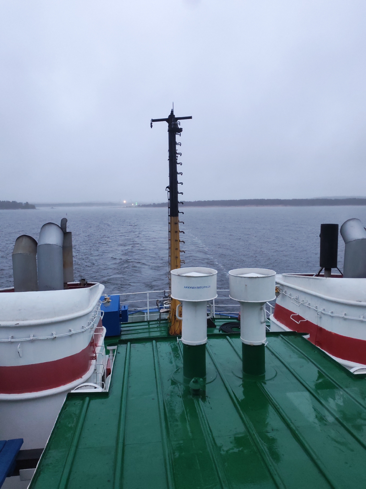 The image depicts a view from the deck of a ferry or boat, likely in the vicinity of Shal'skii, Russia, given the Cyrillic text visible on the white ventilation stacks. The deck is painted green and appears wet, possibly from rain or mist.
In the foreground are two white cylindrical ventilation stacks, labeled with the Cyrillic word "АККУМУЛЯТОРНАЯ" (AKKUMULIATORNAYA), which translates to "battery room" or "accumulator room". To the left and right are lifeboats or similar structures, painted white with a red stripe.
A tall, black mast extends upwards, with various attachments and markings along its length.
In the background, a body of water is visible, with land visible on the far shore. The weather appears overcast and foggy, obscuring the distant details. The overall environment suggests a maritime or riverine setting. There are no people visible in the image.