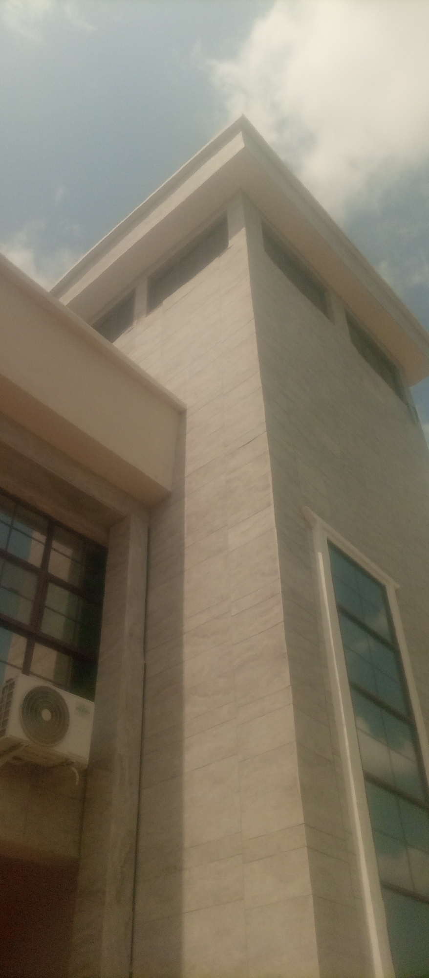 This is a low-angle, slightly tilted, full shot of a modern building in Kuson, Nigeria. The building's exterior walls are clad in light-colored stone with visible horizontal lines. The architecture features clean, sharp angles and a cantilevered overhang on the left side of the frame. Two large windows with dark frames are visible, one on the lower left and another on the right side of the building, reflecting the sky. An air conditioning unit is mounted on the wall below the lower left window. The sky above is bright and partially cloudy.