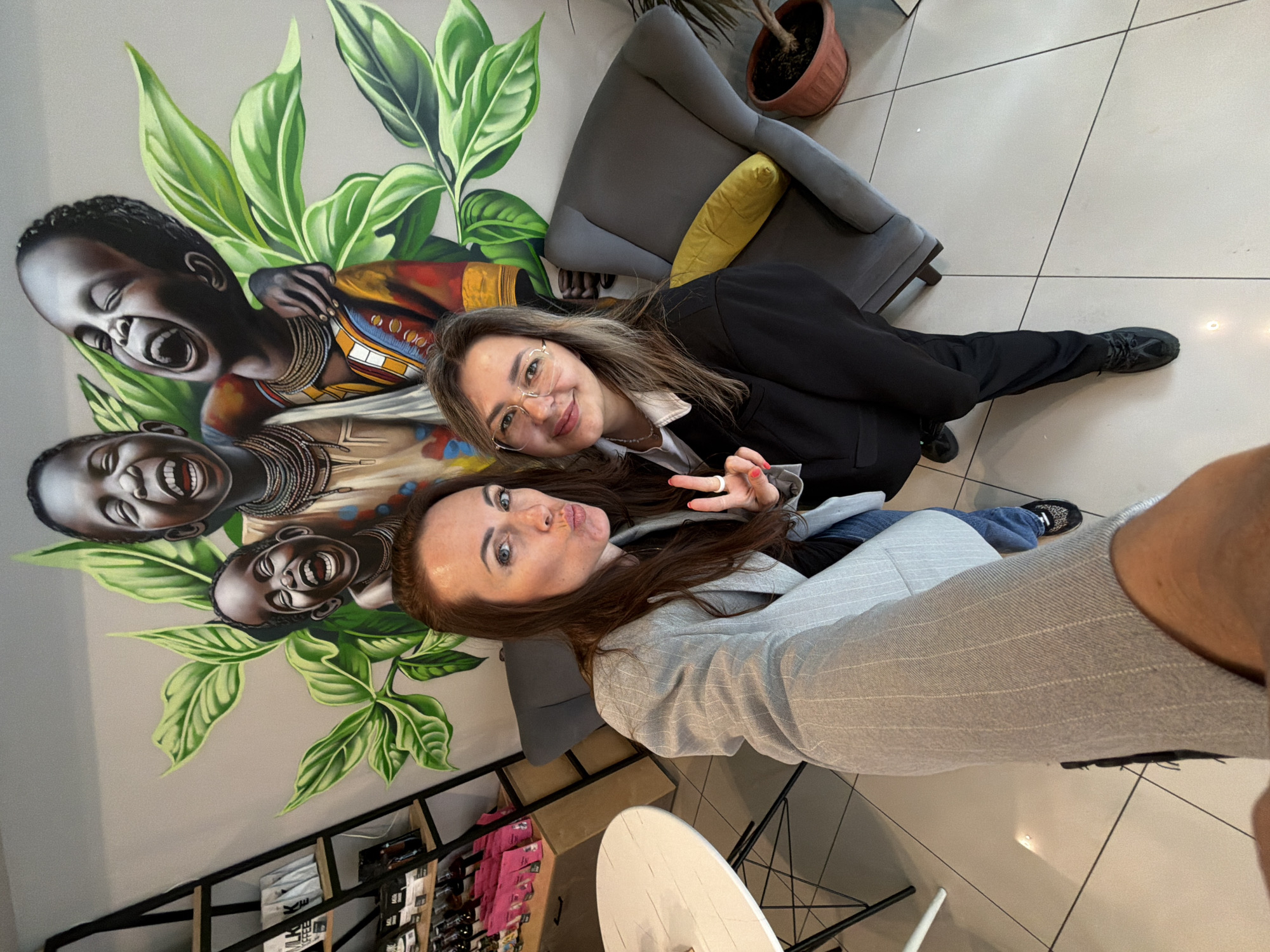 Two women are captured in a selfie taken indoors, likely within a modern commercial establishment such as a cafe or shop in Birobidzhan, Russia. The image has a bright, inviting atmosphere.
The woman on the left, with long reddish-brown hair and blue eyes, is wearing a light gray pinstriped blazer and dark pants, with a partial view of leopard-print sneakers on her right foot. She is playfully pouting her lips and holding up a "peace" sign with her right hand. Beside her, on the right, is another woman with long, light brown hair, wearing glasses, a black blazer over a white collared shirt, black pants, and dark athletic shoes. She is smiling broadly at the camera. Both women appear happy and relaxed, posing for the selfie.
The background features a striking wall mural depicting three dark-skinned children with wide, joyful smiles and open mouths, adorned with elaborate neck jewelry and colorful, traditional-style clothing. They are surrounded by large, vibrant green leaves. Modern furniture is arranged around them, including a gray armchair with a bright yellow cushion, a dark gray chair with a wire base, and a small, round white table. The floor is covered in large, light-colored square tiles with dark grout lines. In the deeper background, a shelving unit with various items is visible, and some packages display indistinct text, with what appears to be Japanese characters on one small item. A potted plant in a terracotta pot adds a natural touch to the scene.