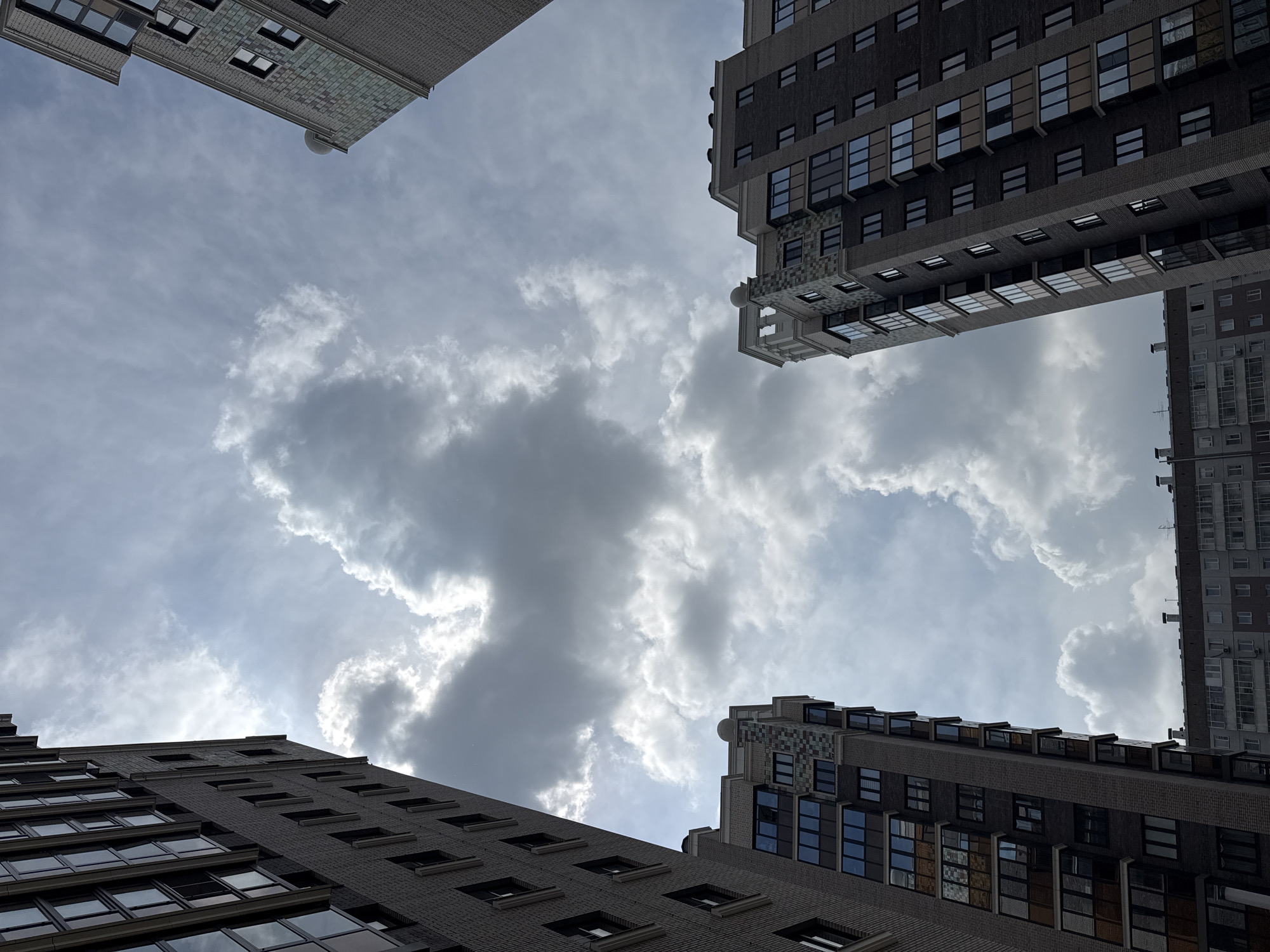 The image shows a low-angle view of several modern apartment buildings against a cloudy sky. The buildings are made of brick and have many windows. Some of the buildings feature decorative elements at the top. The sky is filled with dramatic clouds, with patches of sunlight breaking through. The composition creates a sense of looking up at the imposing structures. The specific location is likely a residential area in the city of Krasnoyarsk, Russia, given the architectural style and common urban landscape. There are no people or visible actions in the image.