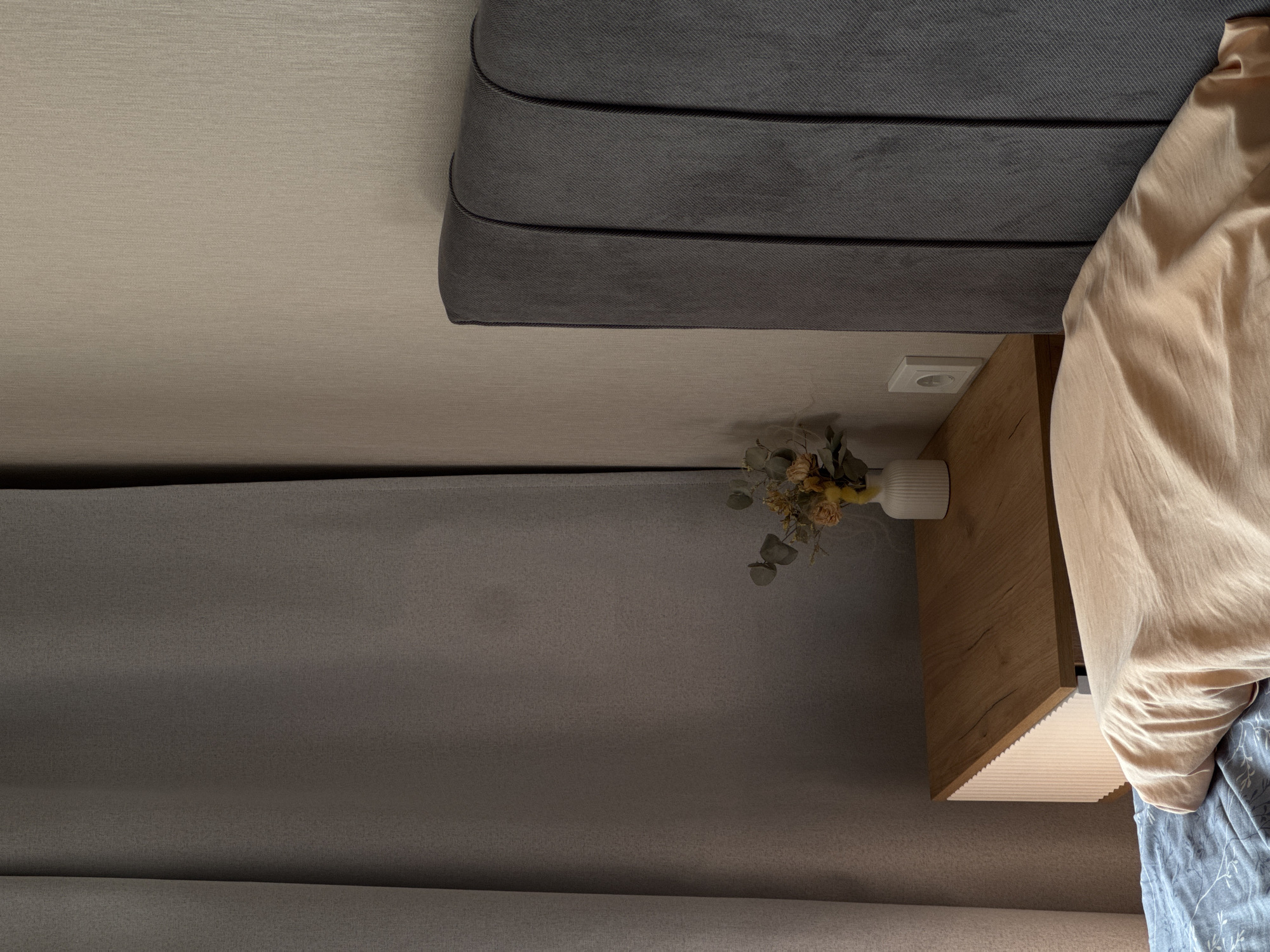A close-up, eye-level shot captures a corner of a bedroom. To the left, a thick, grey curtain hangs, with a subtle texture visible. To the right, a grey upholstered headboard with vertical stitching is partially visible, positioned against a textured beige wall. A small, light wood bedside table sits between the curtain and the headboard. On the table, a white vase holds a small arrangement of dried flowers and greenery. A white electrical outlet is visible on the wall above the bedside table. In the foreground, a bed is partially visible, with a peach-colored pillow and a blue patterned sheet. The scene is lit by soft, natural light coming from the left.