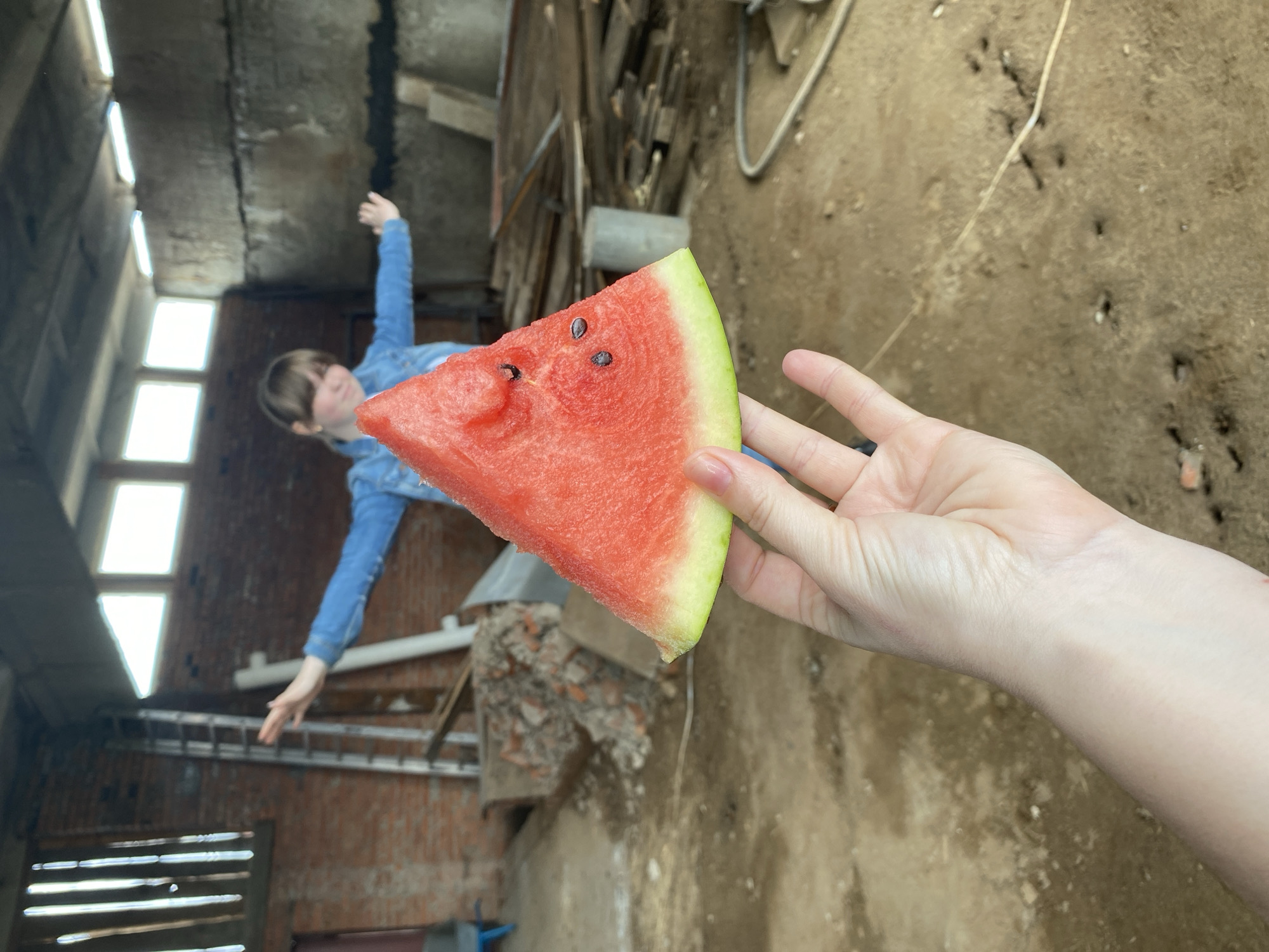 A person holds a watermelon slice in front of a blurry background of a dilapidated building. The watermelon is red with black seeds and a green rind. The person holding it has pale skin. In the background, a young woman in a denim jacket stands with her arms outstretched. The building appears to be in disrepair, with exposed brick and what appears to be damaged or incomplete sections. The picture was taken in Chaikovsky, Russia.
