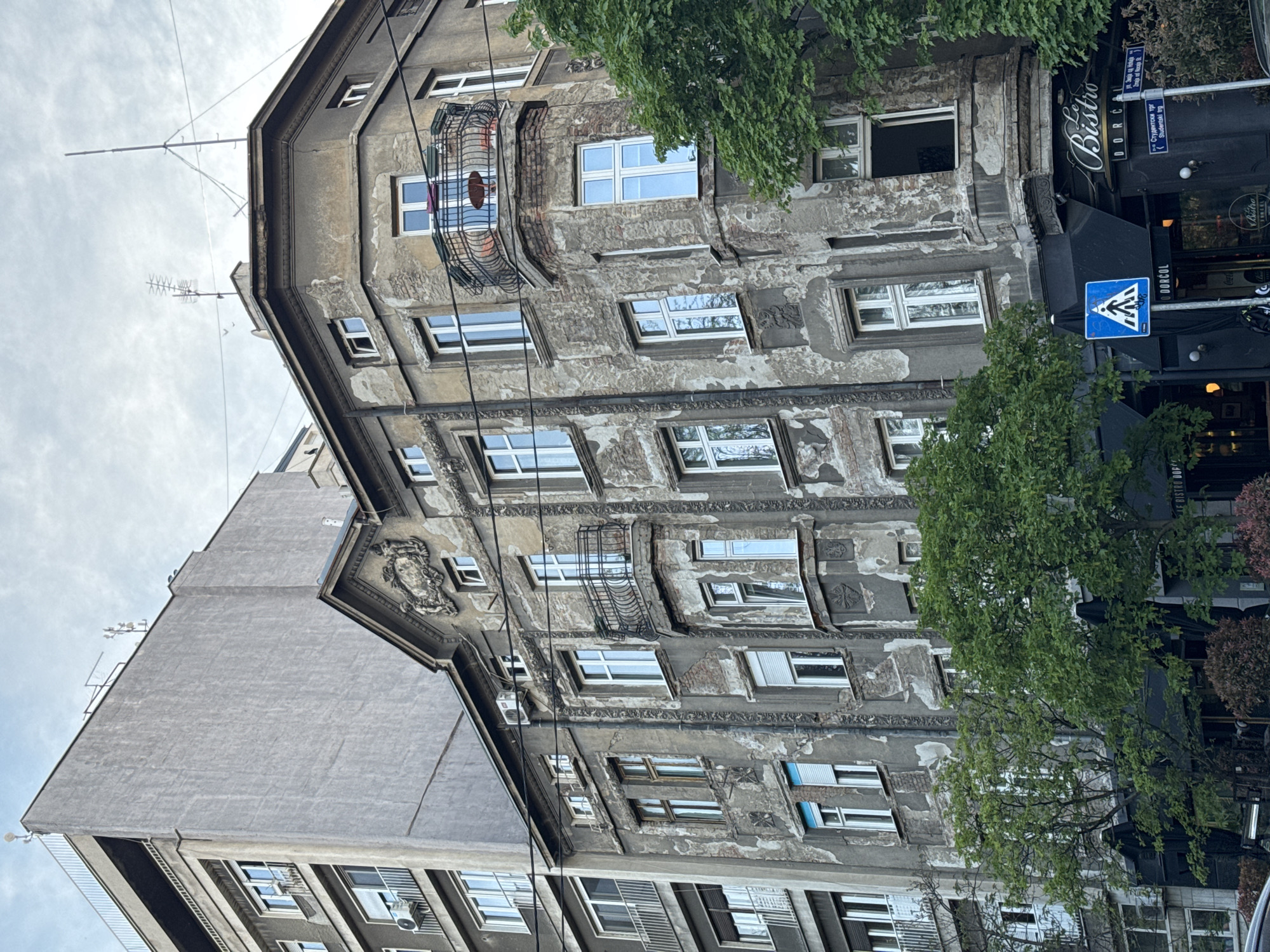 The image depicts a building in Belgrade, Serbia, located at the intersection of Visnjiceva and Studentski Trg streets. The building's facade is weathered and peeling, revealing layers of plaster and brick. It features multiple windows with white frames and at least one ornate balcony with wrought-iron detailing. A modern grey addition is adjacent to the old building. Overhead power lines are visible in the foreground. Street signs and a crosswalk sign are also visible, partially obscured by trees. A restaurant or bistro named "Le Bistro Dorcol" is located on the ground floor of the building.