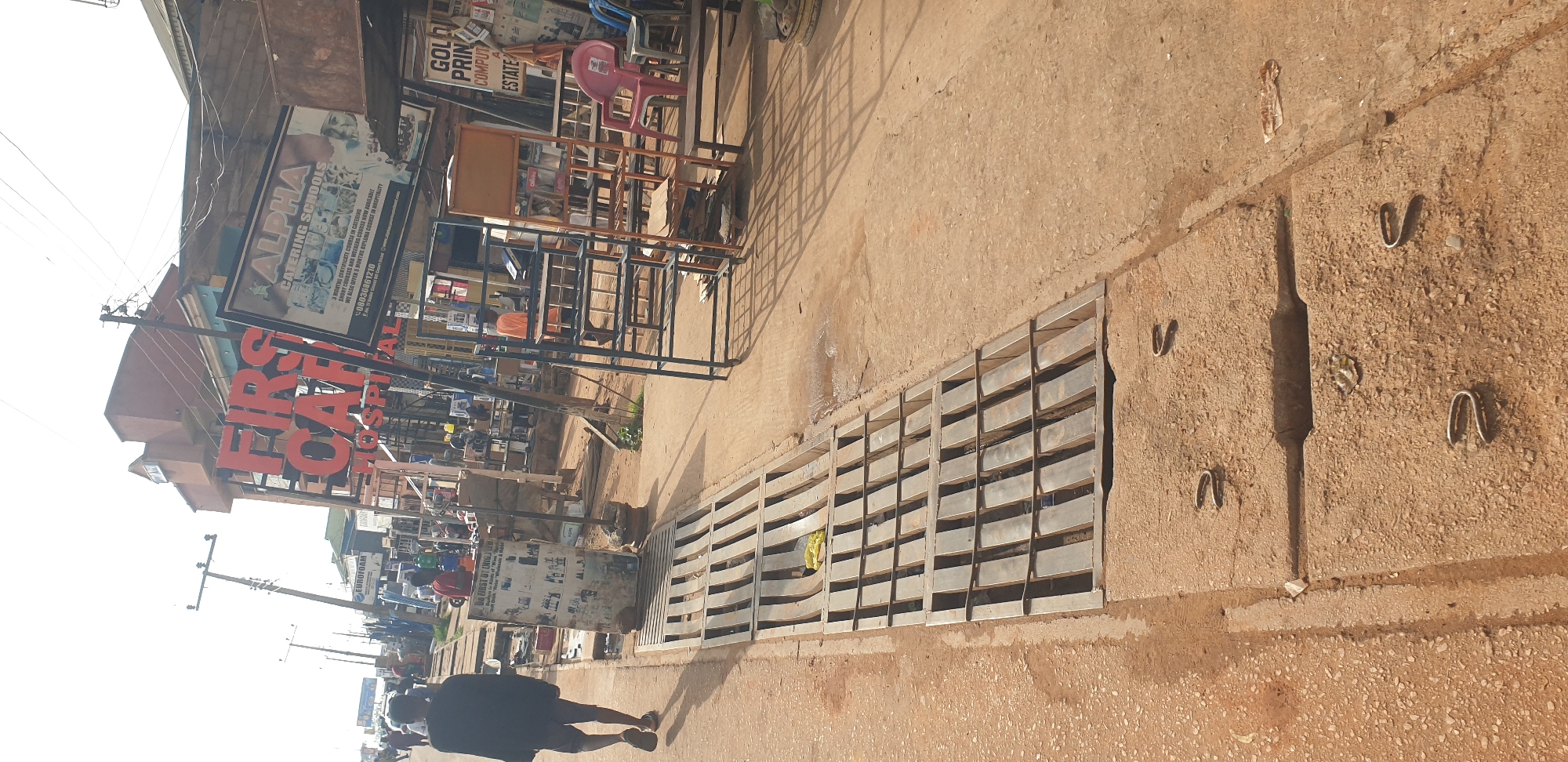The image captures a sunlit street scene in Sabon Tasha, Nigeria, depicting a bustling commercial area. The perspective is at ground level, looking down a paved street or sidewalk lined with businesses.
In the foreground, a person walks away from the camera, dressed in a dark t-shirt and pants, casting a long shadow on the light-colored concrete ground. The pavement features several metal grates, likely storm drains or utility covers, and exposed metal hooks. Bright sunlight and distinct shadows suggest a clear day, possibly mid-morning or late afternoon.
Along the right side of the street, various metal display racks and shelving units are arranged outside shops, possibly for showcasing merchandise, alongside a pink plastic chair.
The left side of the street is lined with buildings housing various businesses. Several prominent signs are visible:
* A large red sign reads "FIRS CA" and "HOSPITAL" (likely "FIRST CAFÉ" or similar).
* Above it, a sign for "ALPHA CATERING SCHOOLS" advertises "3 MONTHS CERTIFICATE COURSE IN CATERING," "SHORT COURSES AND WEEKEND COURSE NOW AVAILABLE," and "5 MONTHS DIPLOMA COURSE IN HOSPITALITY," along with a phone number "08036861210."
* Further down, a sign mentions "GOLD PRIN COMPUT A ESTATE" (likely "GOLD PRINCE COMPUTER ESTATE").
* A poster for "EUROFOAM" is also visible.
Utility poles with numerous overhead wires run along the side of the street. The overall scene depicts a typical commercial thoroughfare with daily activity.