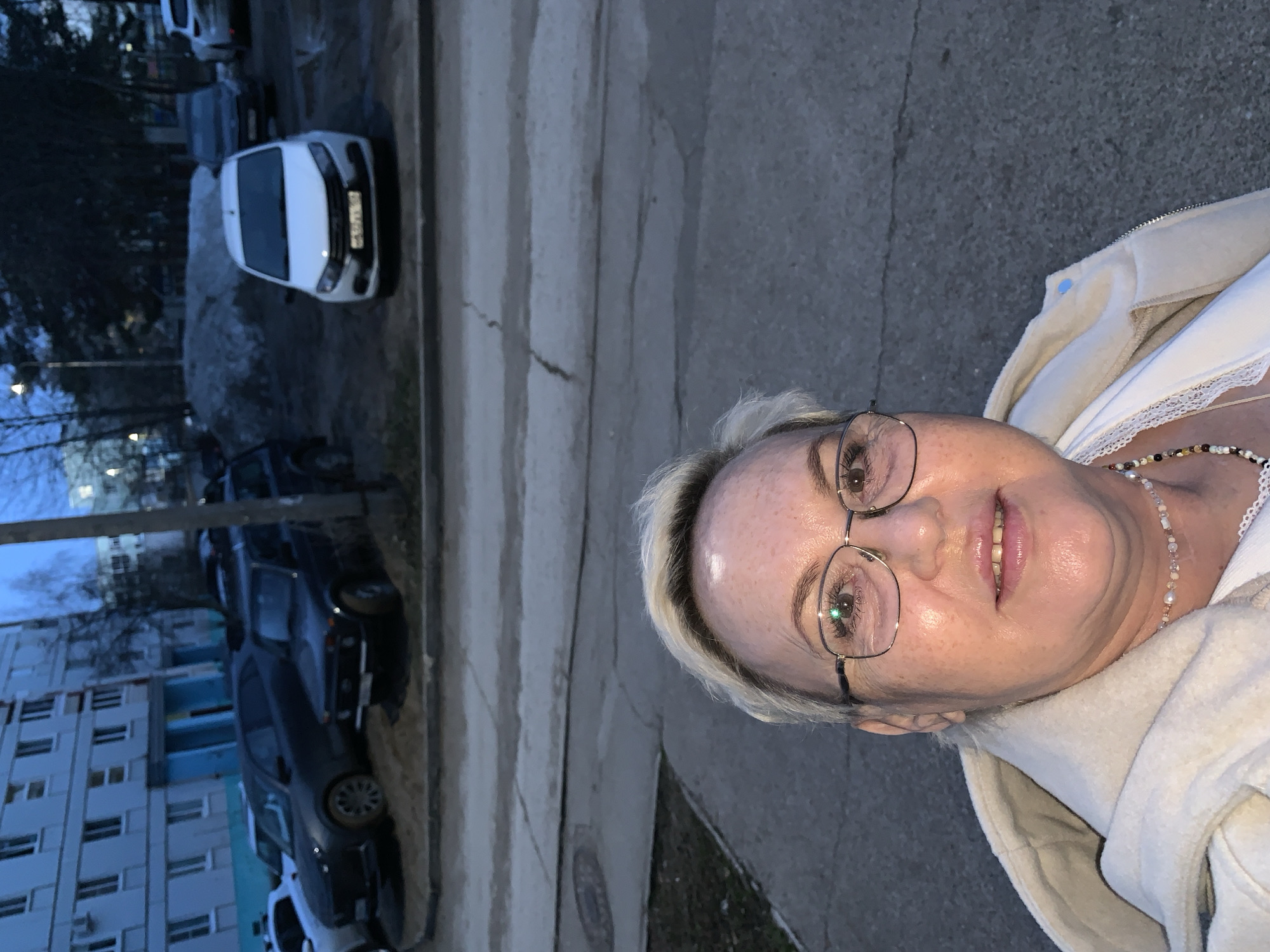 The image is a vertical selfie of an adult woman taken outdoors in Chaikovsky, Russia, during twilight hours. The woman, positioned in the right foreground, has short, light blonde hair and wears glasses. She is smiling faintly, dressed in a light-colored, possibly fleece, jacket over a white top with a lace trim, and a multi-colored beaded necklace.
The background reveals a street scene. A white car, identifiable as a Lada Granta, is parked on the asphalt. Its Russian license plate is partially visible, displaying characters such as "M21XAO59", with "59" indicating the Perm Krai region, where Chaikovsky is located. Further back, a dark-colored sedan, possibly an older Lada model, is also parked. Bare trees with visible branches suggest a cold season, and multi-story residential buildings are seen in the far left background. The ground surface appears damp or possibly icy in places.
The overall lighting is dim, characteristic of dawn or dusk, with a cool blue hue to the sky and environment. The scene depicts a casual moment against the backdrop of a typical Russian residential street.