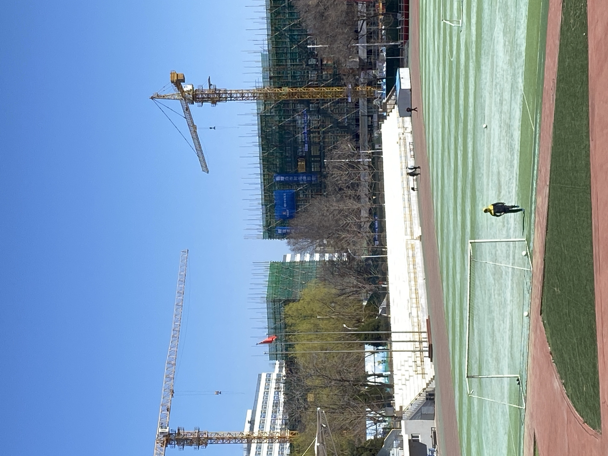 A ground-level outdoor shot, taken during daytime, shows an athletic field and construction sites in the city of Zhongtun, China. The field, oriented vertically, has alternating green and light green stripes, with red running tracks on its edges and white soccer goals. At least one person is visible on the field wearing yellow. A partially constructed building is to the left of the field, with yellow construction cranes and scaffolding visible. In the background, a red flag flies. The sky is light blue.