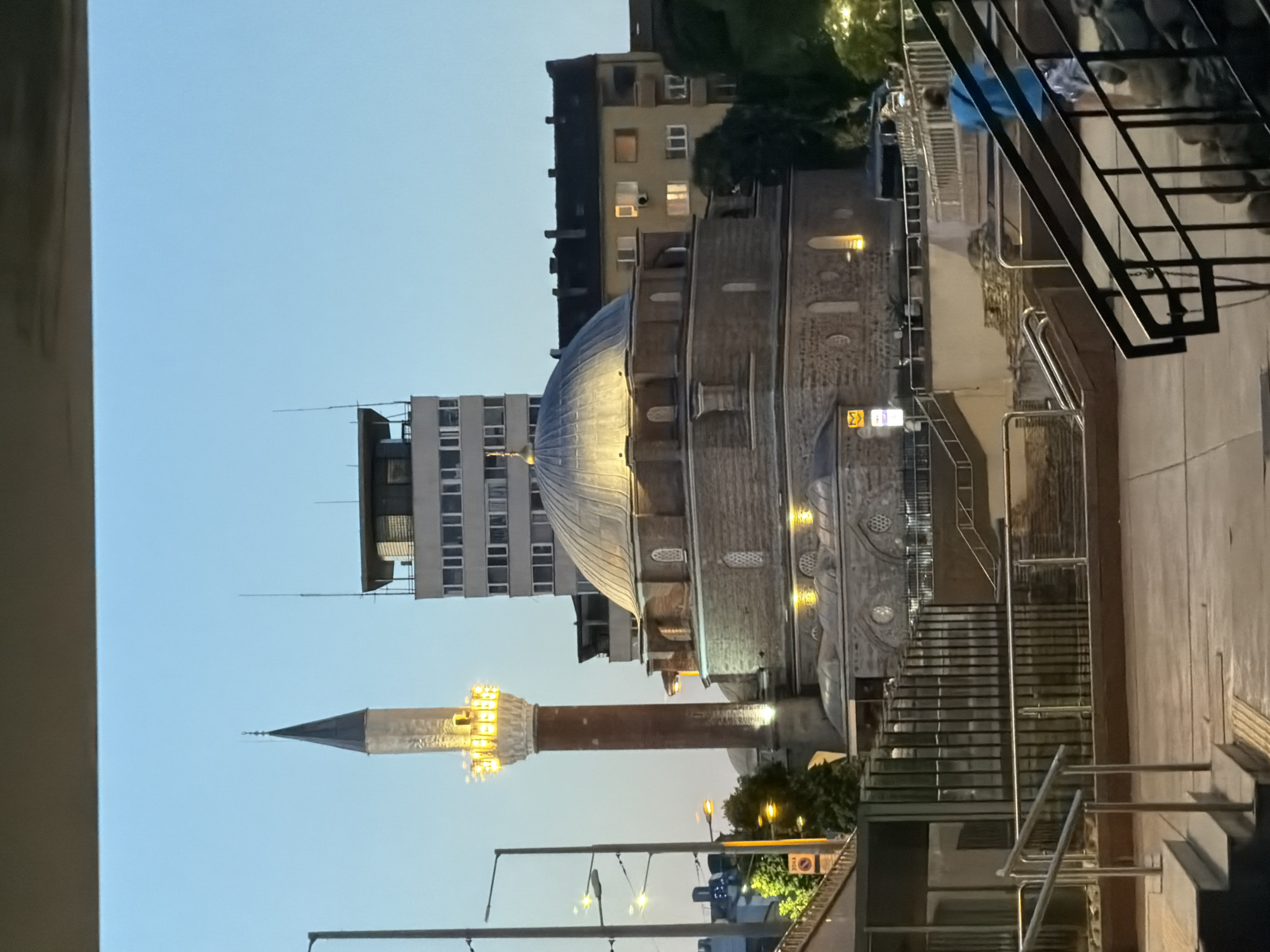 The image depicts the Banya Bashi Mosque in Sofia, Bulgaria, at dusk. The mosque's dome and minaret are illuminated, while the sky is a pale blue, indicating twilight. A tall, modern building with multiple windows stands behind the mosque. In the foreground, a person wearing a blue t-shirt and patterned shorts walks away from the camera down a set of stairs. Metal railings and a concrete walkway are visible in the foreground, and there are some low walls and foliage on the sides. The lighting suggests an urban environment transitioning into evening.