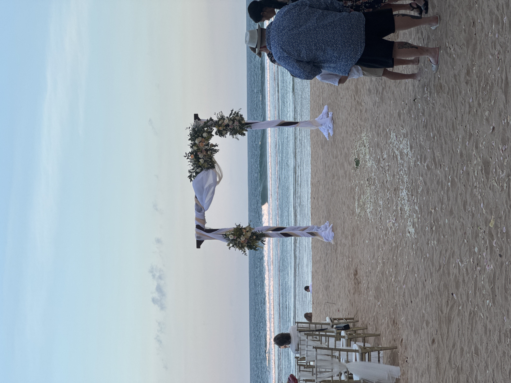 A beach wedding ceremony is set up on the sand in Kuta Selatan, Indonesia. A wooden arch adorned with white drapery and floral arrangements stands as the focal point, framing the ocean and sky. Rows of empty chairs are arranged on the left side of the sand, indicating where guests would be seated. Several people are visible, including a woman with dark hair in a patterned blue shirt and black shorts standing with her back to the camera, facing the ocean. Another person, wearing a white hat, is seen from behind to the right of the arch, also looking towards the sea. Children are also present, and a few individuals are seated on chairs in the background. The sand is scattered with white flower petals, and the ocean waves gently roll onto the shore under a clear sky with a hint of clouds.