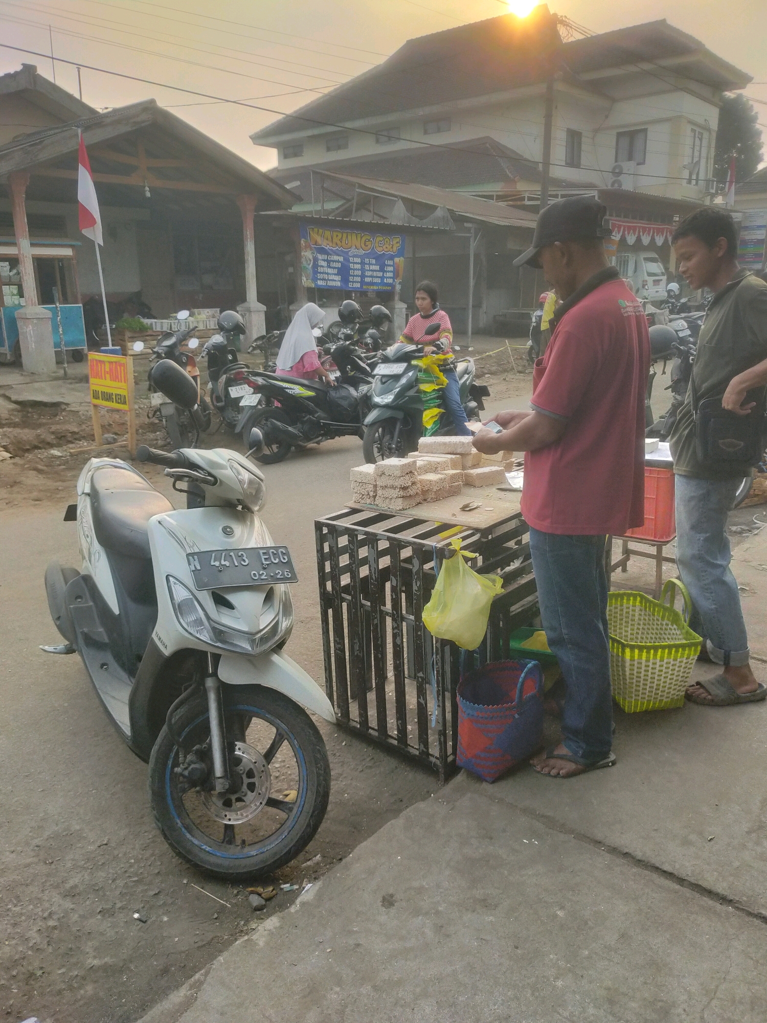 A street scene in Bululawang, Indonesia, features a man in a red shirt and black cap standing at a food stall, arranging stacks of what appear to be crackers. He is interacting with another person in a green shirt and jeans to his right. A white scooter is parked in the foreground. The background shows a row of buildings with awnings, some with Indonesian flags, and several motorcycles parked in front of them. A sign with Indonesian text, likely for a warung (small shop), is visible. The lighting suggests it is late afternoon or early evening, with the sun low in the sky. The street is paved, and there are various baskets and bags around the stall.
