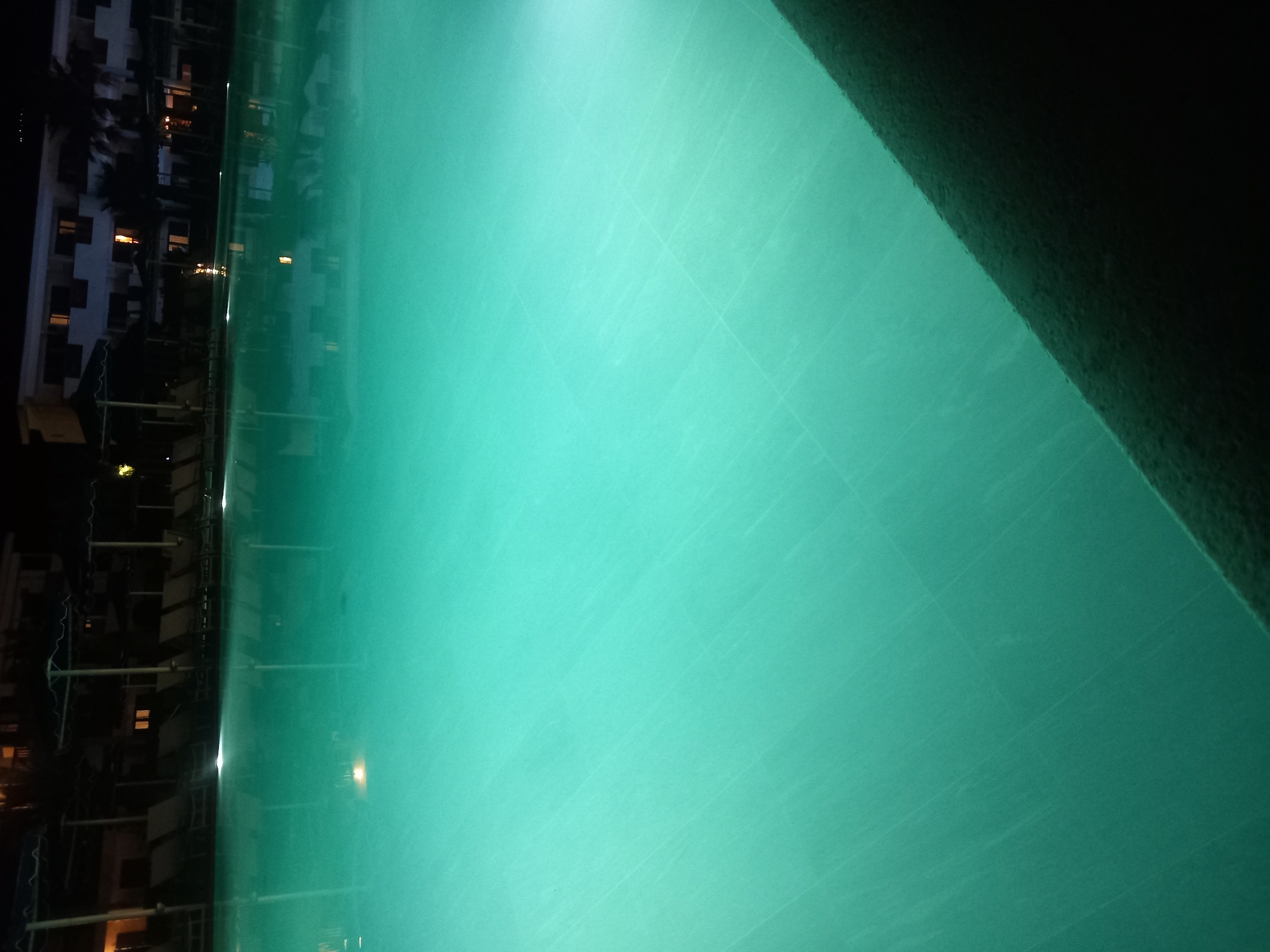 The image depicts an outdoor swimming pool at night, illuminated by underwater lights which cast a vibrant turquoise glow. The water is calm with slight ripples. The edge of the pool, a dark, textured surface, occupies the lower right portion of the frame. In the background, a building with visible windows that are lit from within, along with several lounge chairs and umbrellas, suggest a hotel or resort setting. The presence of palm trees further indicates a warm climate. The overall scene is serene and tranquil, characteristic of a nighttime ambiance at a resort. The location is identified as Kos, Greece.