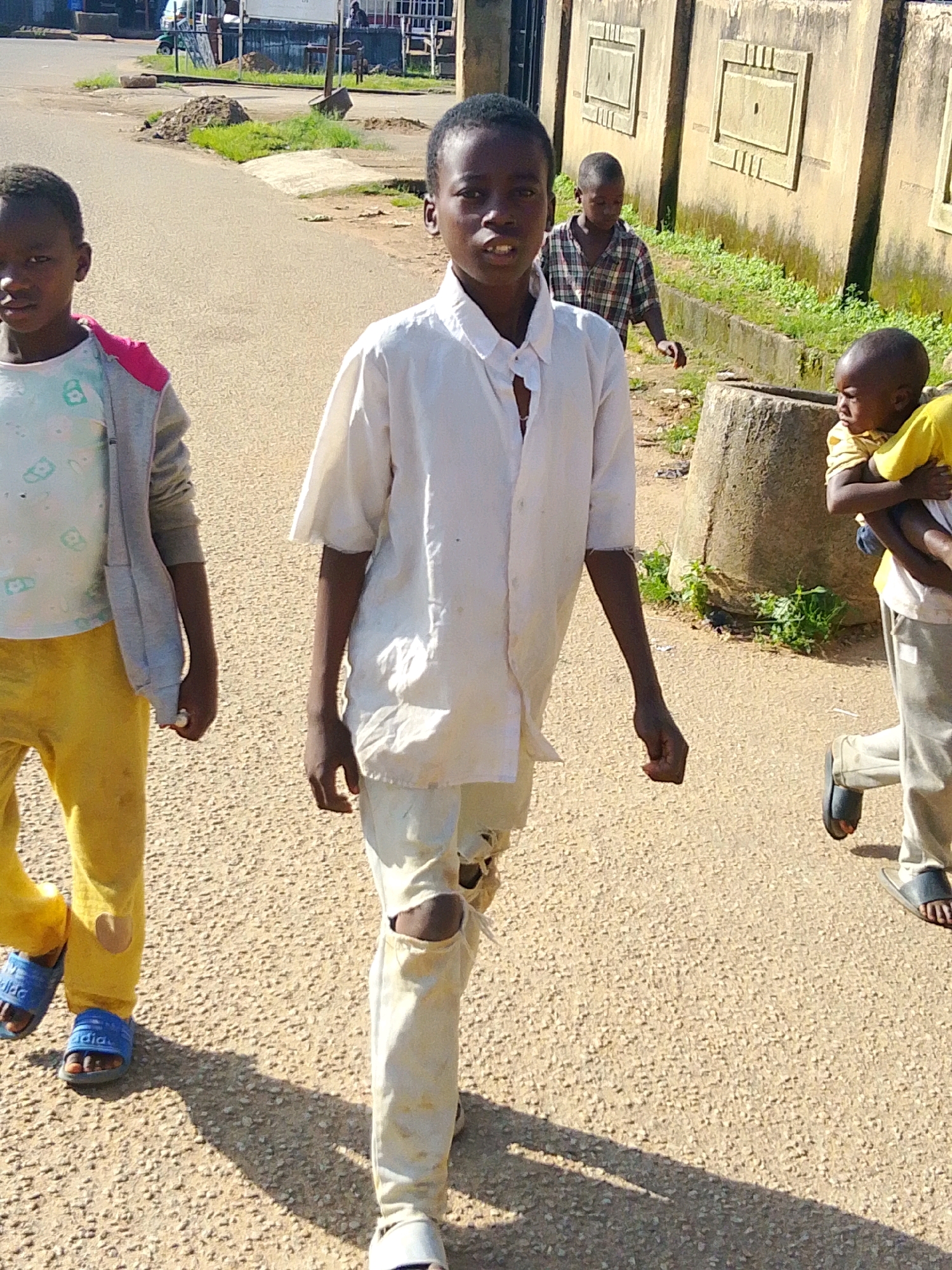 In Jos, Nigeria, four young boys walk along a dusty, paved road under bright sunlight, casting distinct shadows. The boy closest to the camera, positioned in the center, wears a slightly rumpled, off-white button-down shirt and torn, pale trousers. To his left, another boy in a grey hooded jacket over a t-shirt and bright yellow trousers walks with his hands loosely at his sides. Further back and to the right, a boy in a plaid shirt is visible, his gaze directed downwards. On the far right, a boy in a yellow shirt carries a much younger child who is also wearing yellow. The background features a low, textured, tan-colored wall with rectangular decorative elements, bordering a strip of green vegetation. A large, cylindrical, weathered object, possibly a wellhead or concrete post, stands near the wall. The road surface is a light brown, granular texture, indicative of aggregate. The overall scene depicts an everyday moment of children in an outdoor urban environment in Jos.