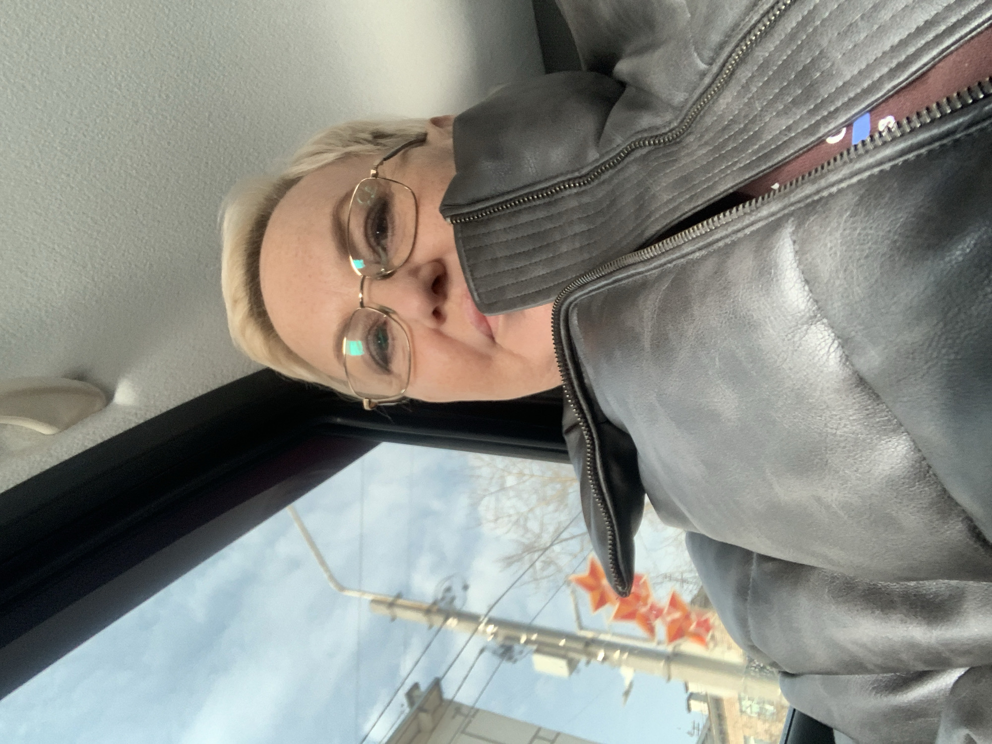 An individual, likely a female adult with short blonde hair and gold-rimmed glasses, is captured in a close-up image, appearing to be a selfie. She is seated inside a vehicle, evidenced by the light-colored, textured ceiling and dark window frame visible above and to her left. She is wearing a dark grey, possibly faux leather, padded jacket with a partially unzipped front, revealing a maroon garment underneath. Her expression is neutral, and her gaze is directed towards the camera.
Through the vehicle's window, an exterior street scene in the city of Chaikovsky, Russia, is visible under a blue sky with scattered clouds. A light-colored utility pole featuring multiple cross-arms and power lines is prominent on the left. Suspended below the utility pole are a series of three orange, five-pointed star decorations. In the background, beyond the pole and decorations, bare tree branches and sections of light-colored, multi-story buildings with visible windows are discernible. The scene is illuminated by natural daylight.