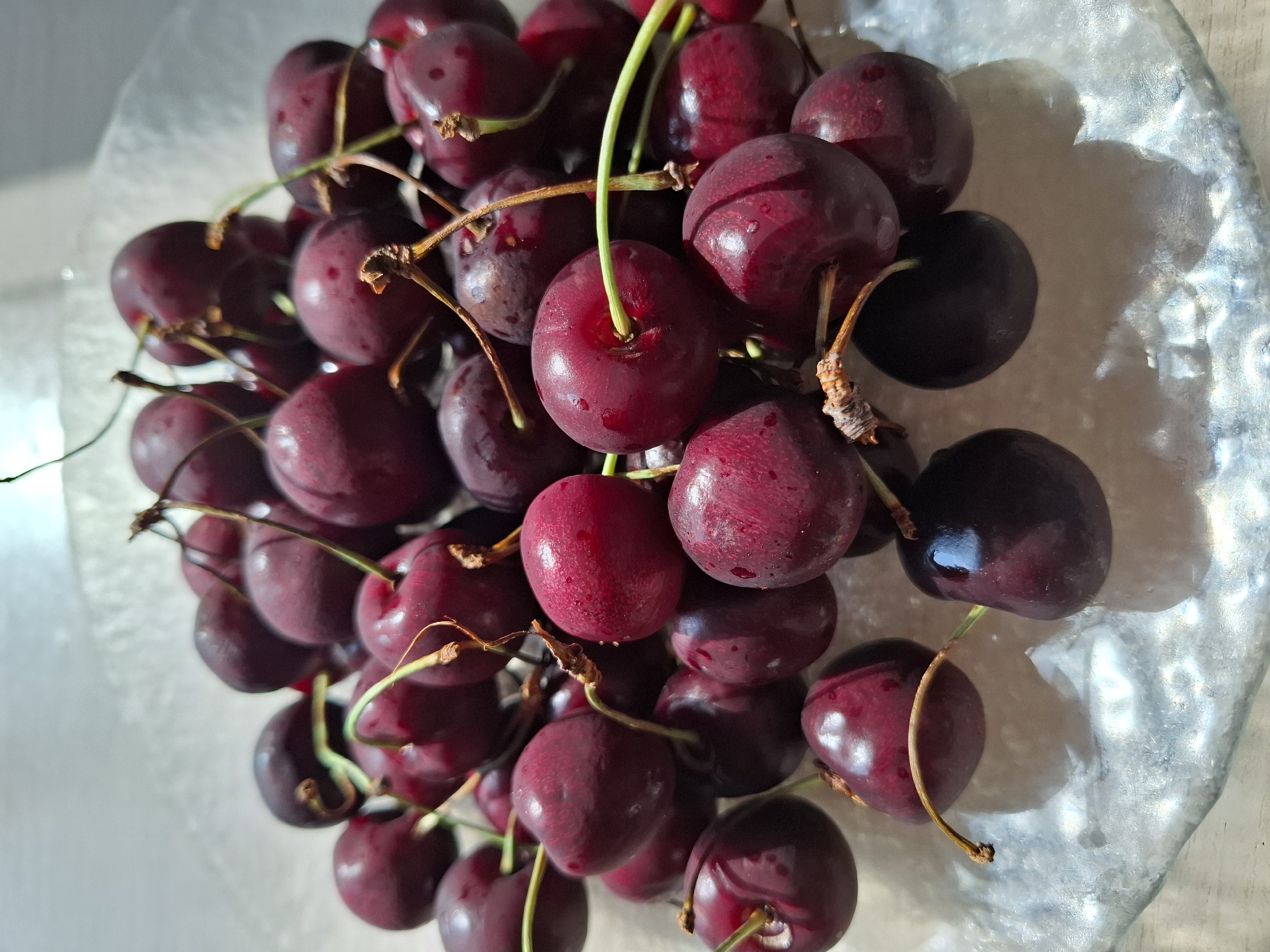 A close-up, high-angle shot shows a pile of dark red cherries on a textured, translucent glass plate. The plate rests on a light-colored, textured surface, possibly a table, with light casting shadows and highlights. The cherries are plump and glossy, some with visible water droplets. The stems are thin and brown, with a few green stems also present, intermingled among the fruit. The lighting suggests an indoor setting, possibly near a window, as evidenced by the strong highlights and shadows. The scene is devoid of any people or human activity. No specific location within Tyumen, Russia, can be identified from the visual information provided.
