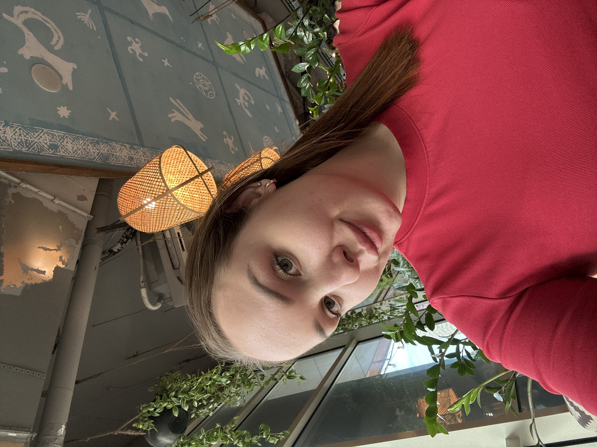 A medium shot angled to the left shows a woman with fair skin and brown hair, wearing a red top. The background features a wall with cartoonish white figures on a blue background, a wicker-style lamp, and greenery. The woman's face is in focus, revealing her eyes and nose. Outside the window, a glimpse of the cityscape of Izhevsk, Russia can be seen.