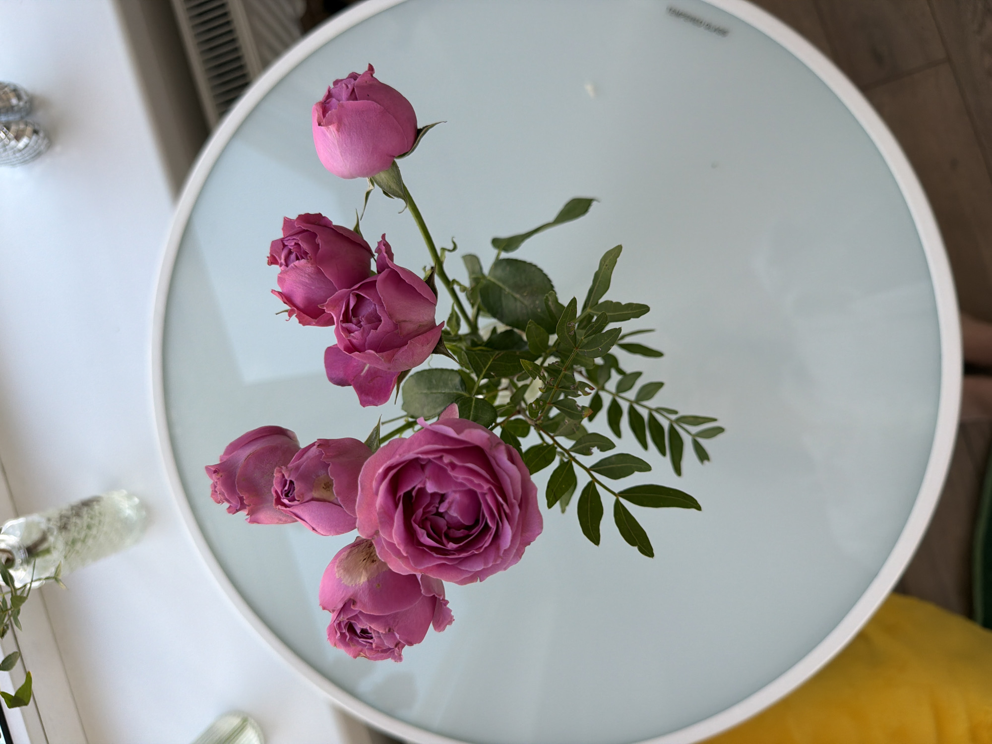 A top-down, high-angle shot shows a bouquet of roses arranged on a round table with a white frame and a light blue, possibly glass, surface. The roses are various shades of pink and purple, with some buds closed and others fully open. Green leaves are interspersed among the roses. In the upper left corner of the image, there are disco balls and a clear glass vase. The table is partially positioned near a window with a white frame. The location appears to be indoors, possibly in Unknown, Unknown.