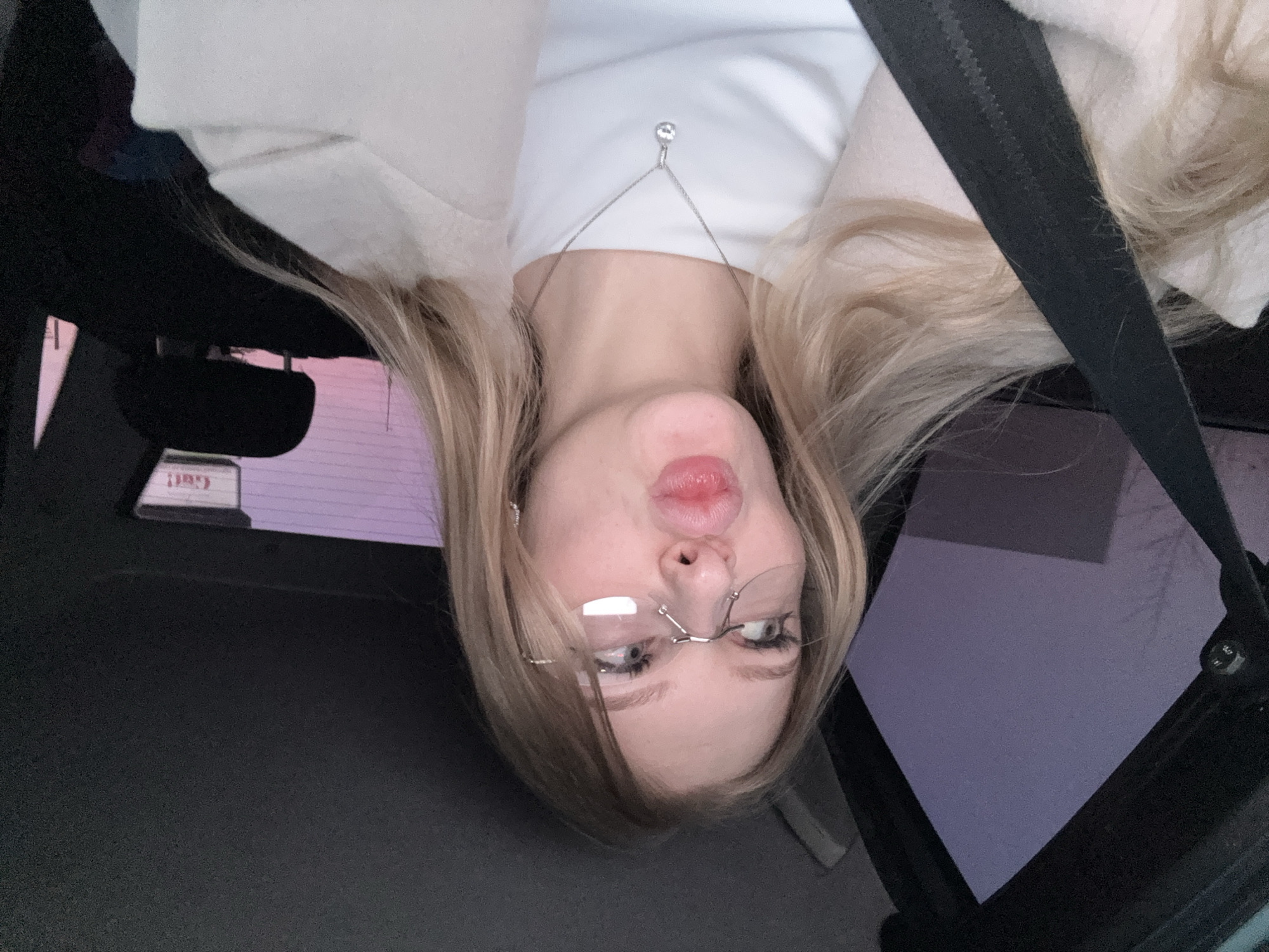 A blonde woman wearing glasses is taking a selfie inside a car, facing the camera. She has a white t-shirt and a light-colored jacket on, with a silver necklace visible. A seatbelt is diagonally across her. The rearview mirror and the side window of the car are visible. The image is rotated such that the woman appears upside down. There is no indication of Perm, Russia in the image.