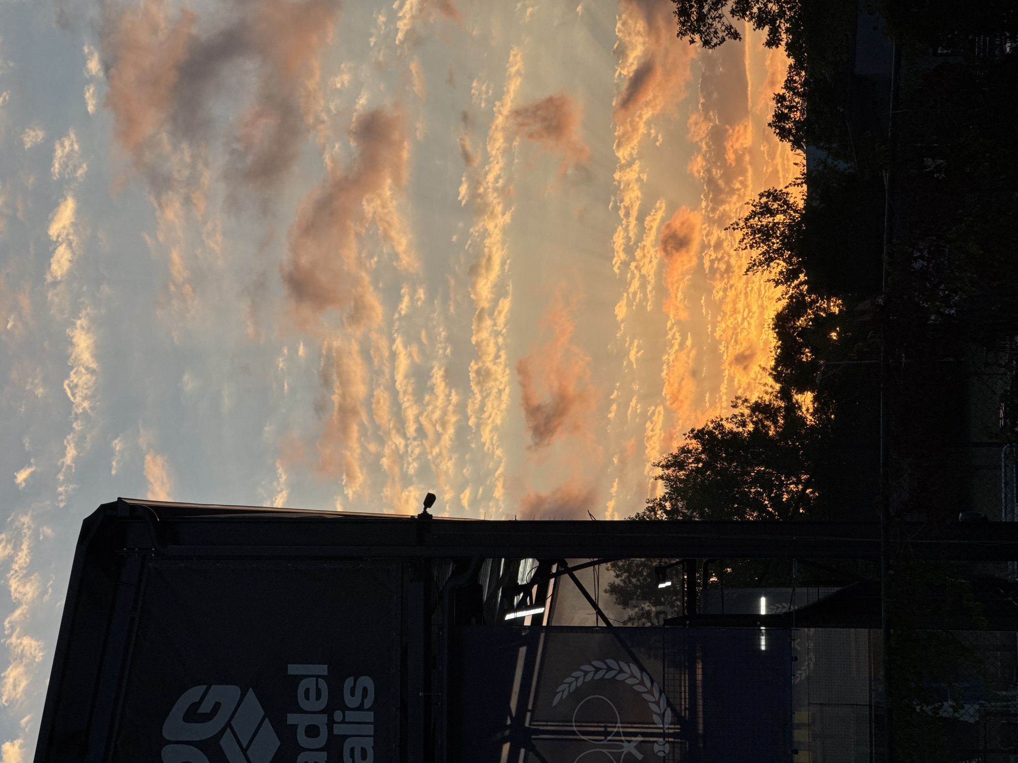 A wide shot captures a dramatic sunset over the city of Tsarevo, Bulgaria. The sky is a vibrant spectacle of orange, yellow, and pink clouds, illuminated by the setting sun. In the foreground, a large, dark structure with the partially visible logo "ad el alis" dominates the left side of the frame. A security camera is mounted on the side of this structure. Below, dark silhouettes of trees and a fence are visible. The scene is devoid of any people or ongoing activities.