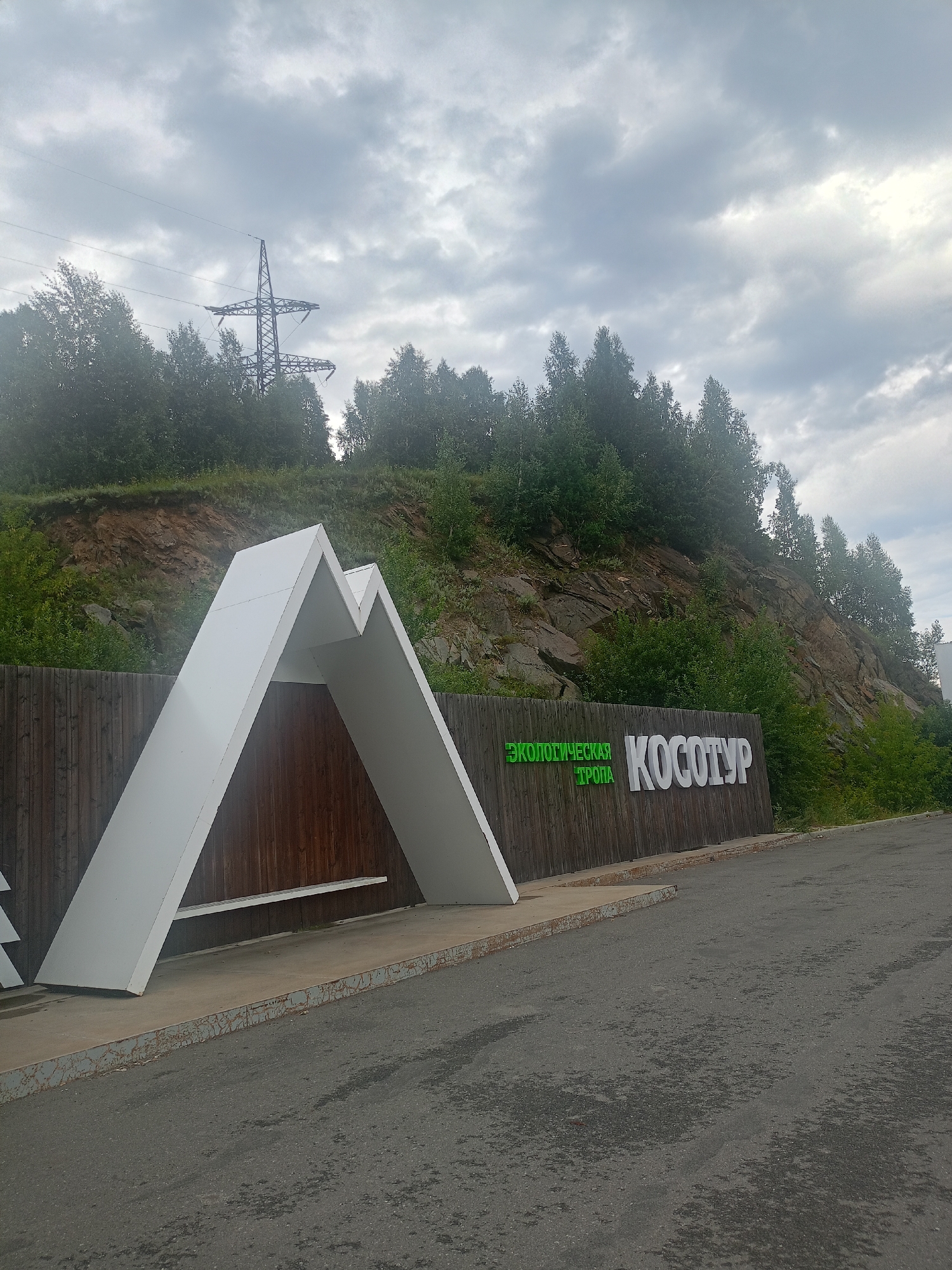 The image shows an outdoor scene with a sign that reads "ЭКОЛОГИЧЕСКАЯ ТРОПА КОСОТУР" in Cyrillic. The sign is made of wood and has large white letters spelling out "КОСОТУР" and green letters spelling out "ЭКОЛОГИЧЕСКАЯ ТРОПА". To the left of the sign is a large white structure shaped like a mountain peak, with wooden paneling behind it. Behind the sign and structure is a rocky hillside covered with dense green trees. The sky is overcast with gray clouds. In the background, a power line pylon is visible above the trees. The foreground shows a paved road or parking area. The location appears to be an entrance to an ecological trail. Based on the sign's text, this is likely the Kosotur Ecological Trail in Zlatoust, Russia. There are no people or animals visible in the image.