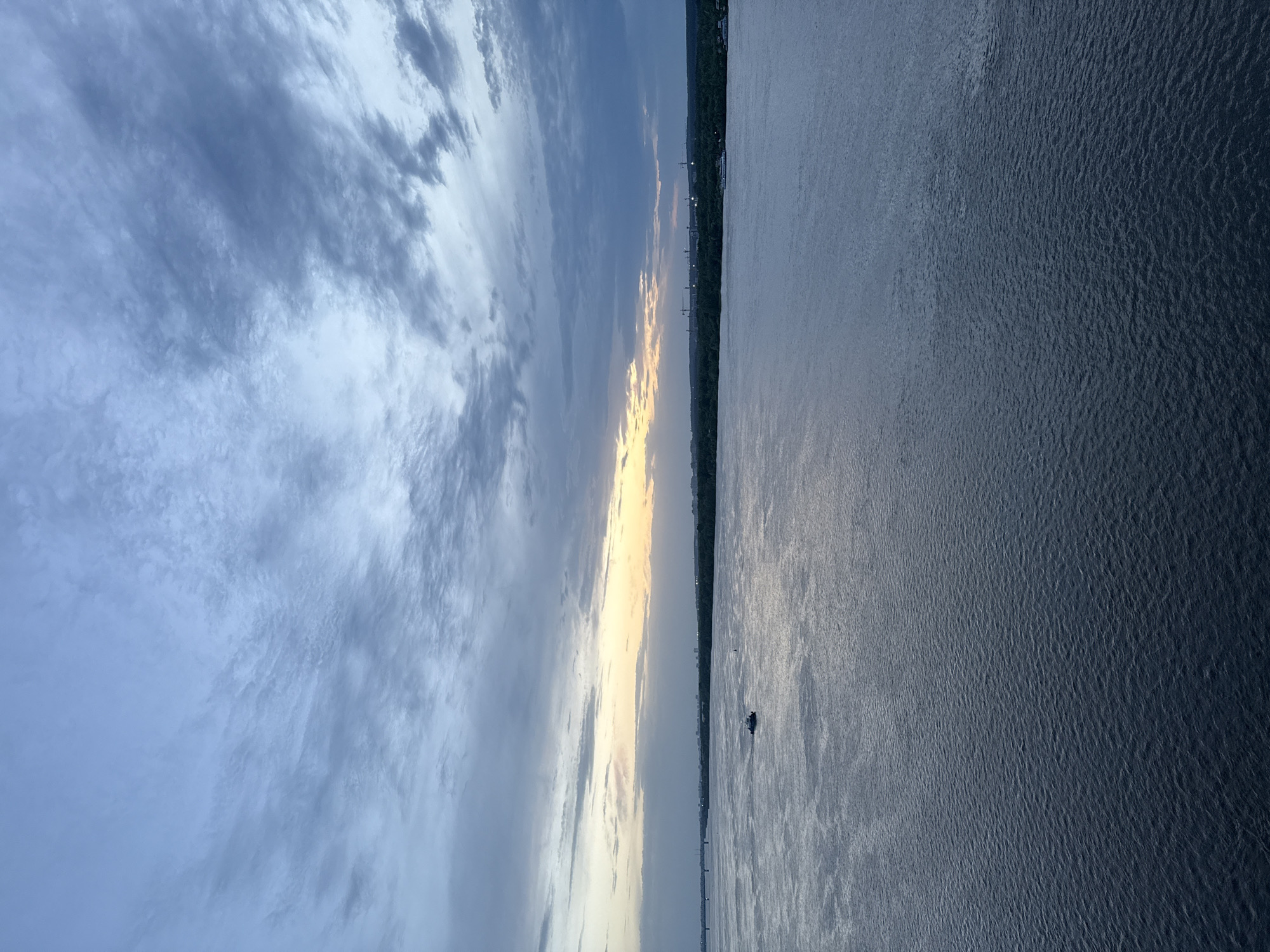 The image depicts a body of water, likely the Kama River in Perm, Russia, as evidenced by the landscape and river's scale. The sky is overcast with clouds, tinged with colors suggesting either sunrise or sunset. The water is dark, reflecting the sky, with slight ripples on the surface. A thin strip of land is visible on the horizon, dotted with artificial light sources and structures, possibly indicating urban development. In the foreground, a small watercraft, possibly a boat, can be seen on the water.