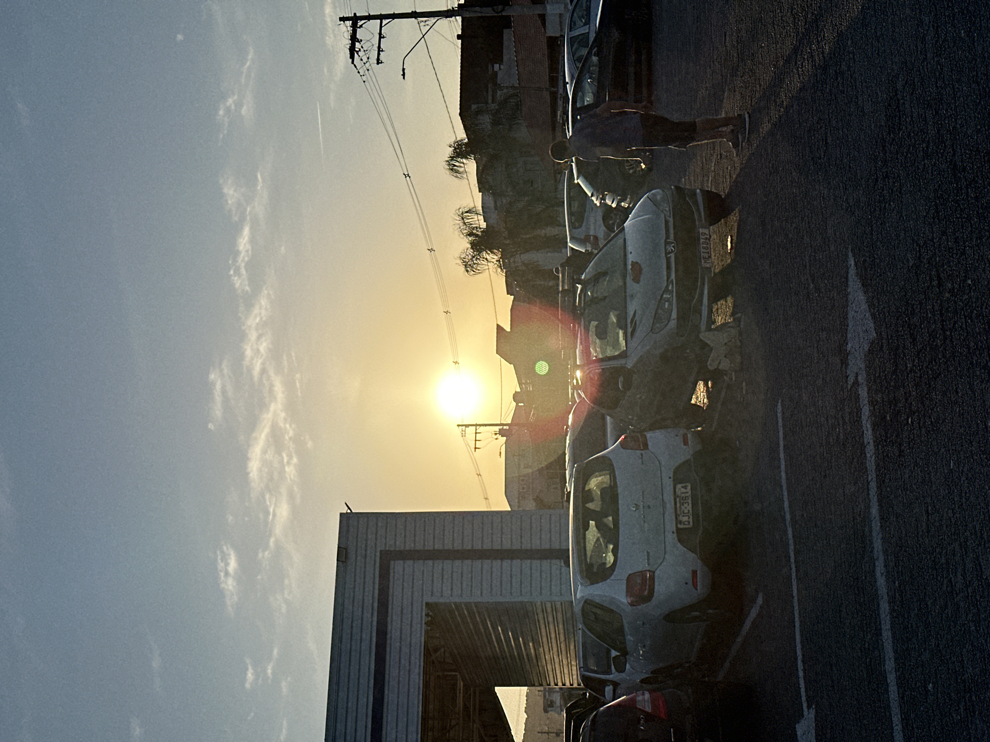 The image depicts a parking lot scene at sunset in Florianópolis, Brazil. Parked cars are aligned, with license plates visible on a few. A person walks past the cars near the asphalt edge of the lot marked with a white arrow. Overhead power lines stretch across the frame, silhouetted against the bright sun. Buildings border the parking area, with a metallic structure visible on the left. The sky is a gradient of blue to orange where it meets the setting sun, with scattered clouds.