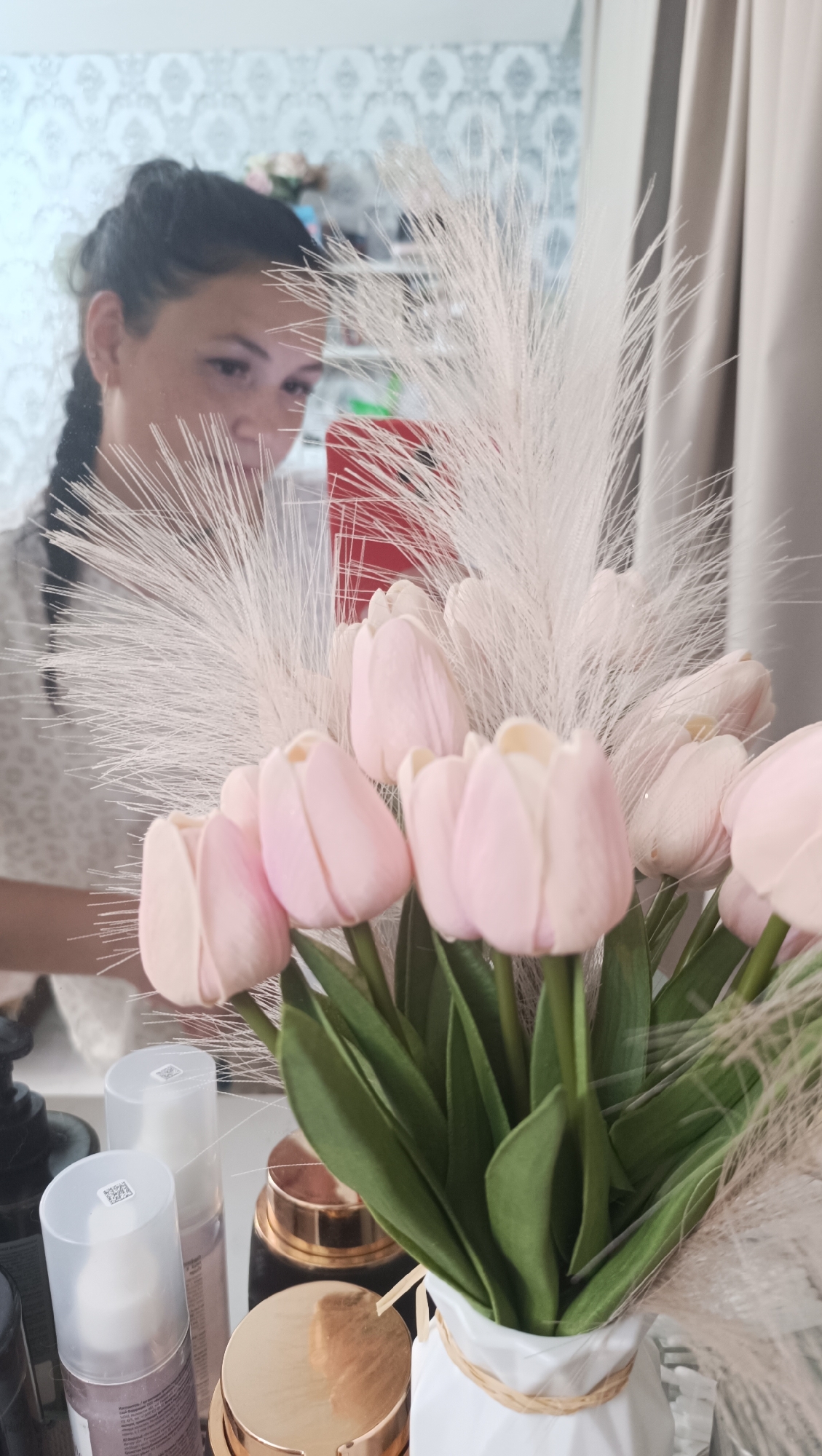 A reflection in a mirror shows a woman with dark hair in a braid, wearing a white patterned shirt, looking at the camera and smiling. She is holding a red mobile phone. In the foreground, a bouquet of light pink tulips and fluffy white plumes is displayed in a white vase tied with twine. Several cosmetic bottles and jars, one with a gold lid, are arranged on a surface in front of the vase. The background of the reflection reveals a patterned wallpaper and what appears to be shelves with items. The overall scene suggests a personal dressing area or vanity. The image does not contain any information about the location in Chaikovsky, Russia.