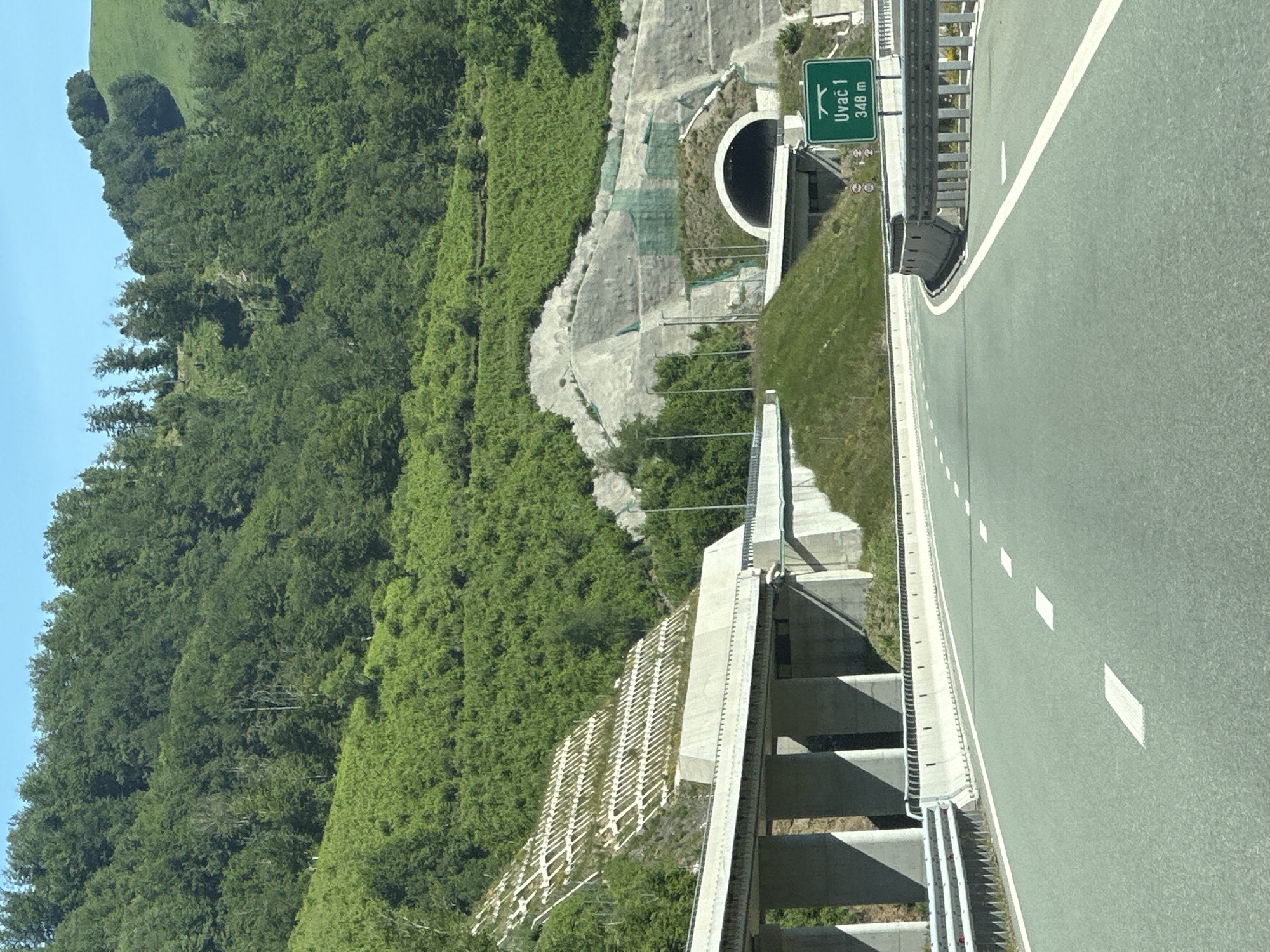 This is a photograph of a highway passing through a mountainous region in Klopot, Montenegro. The highway is paved with a light green asphalt and has white lane markings. On the left side of the highway, there is an elevated overpass supported by concrete pillars, and further up the slope, a reinforced retaining wall with a terraced pattern. On the right side of the highway, there is a tunnel entrance carved into the mountainside, with a green sign indicating "Uvac 1 348 m". The surrounding environment is dominated by lush green vegetation covering the steep slopes of the mountains. The sky is clear and blue. There are no people visible in the image.