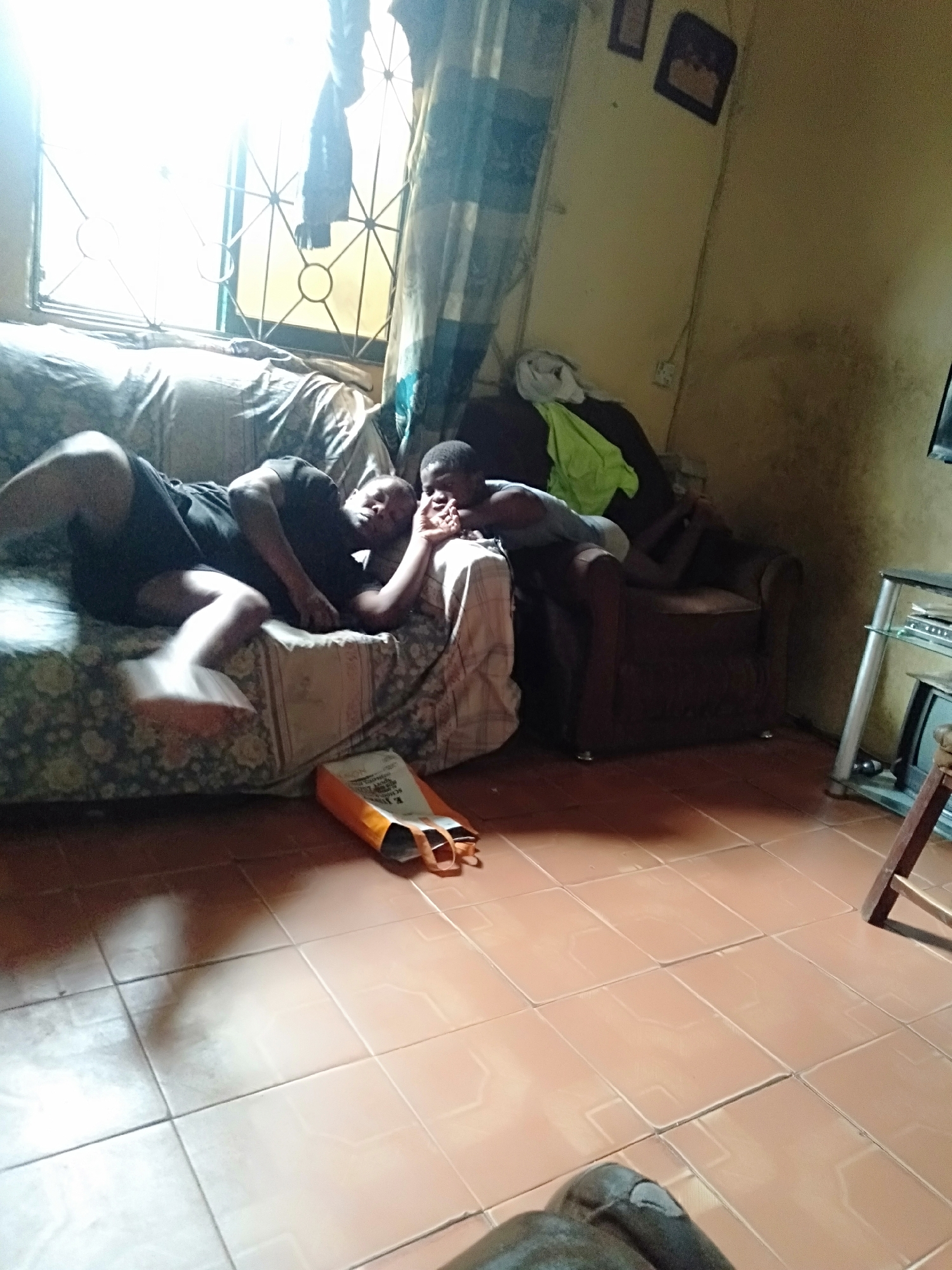 A room in Shangisha, Nigeria, displays two males, one lying on a floral-patterned couch, dressed in dark clothing. The other male sits on a brown armchair, also clothed. A window with bars and sheer curtains is visible behind the couch, letting in daylight. An orange bag rests on the tiled floor next to the couch. A television stand is to the right of the armchair. The walls are yellowish. A pair of framed pictures hangs on the wall behind the armchair.