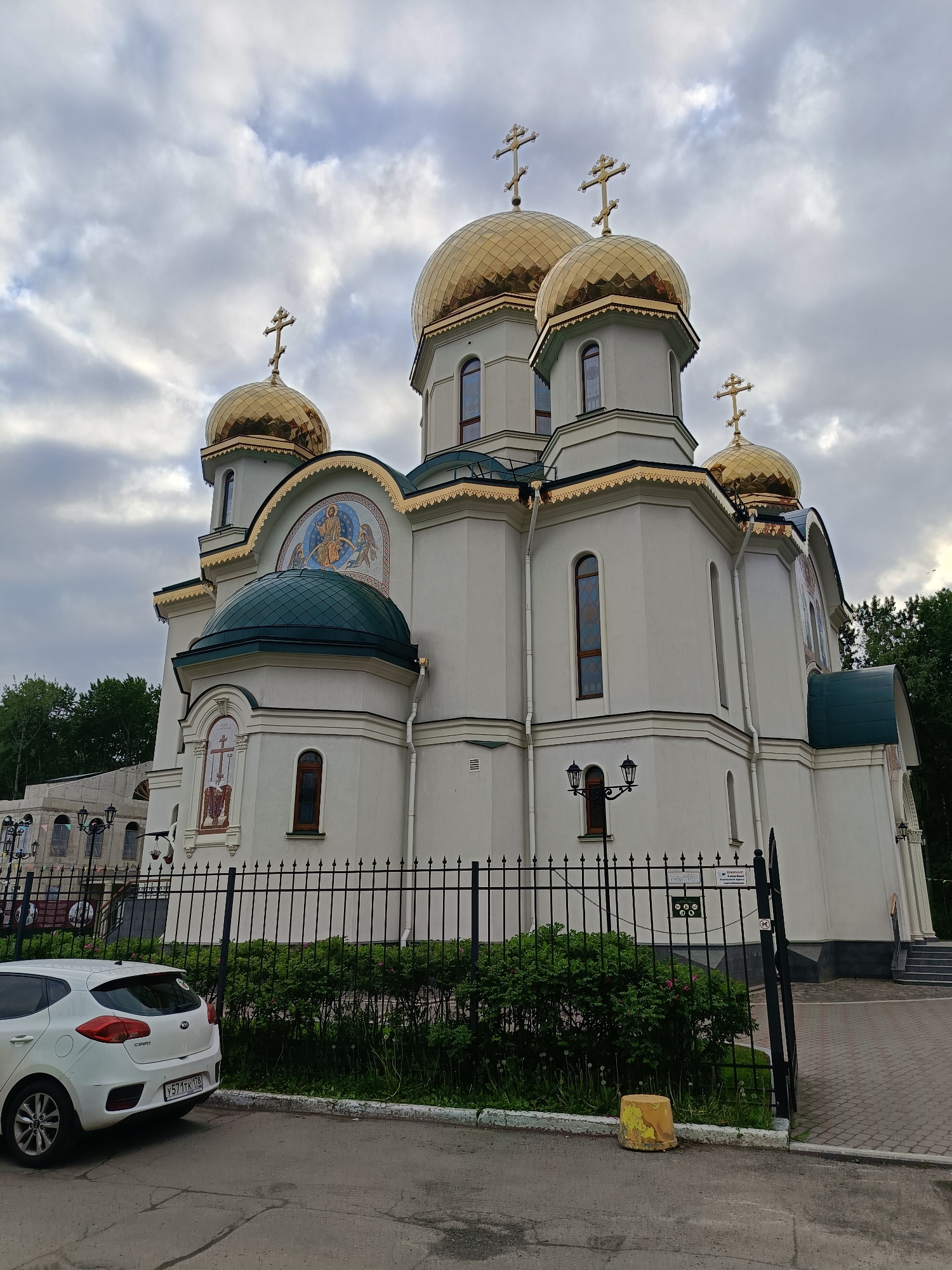 The image shows the Holy Trinity Church (Свято-Троицкий собор) located in Saint Petersburg, Russia. The Orthodox church is characterized by its five golden domes, each topped with a cross. The building's facade is predominantly white, with green accents on some of the roofs. A black metal fence surrounds the property, with some greenery behind it. A white car is parked on the street in front of the fence. The sky is overcast with cloudy weather.