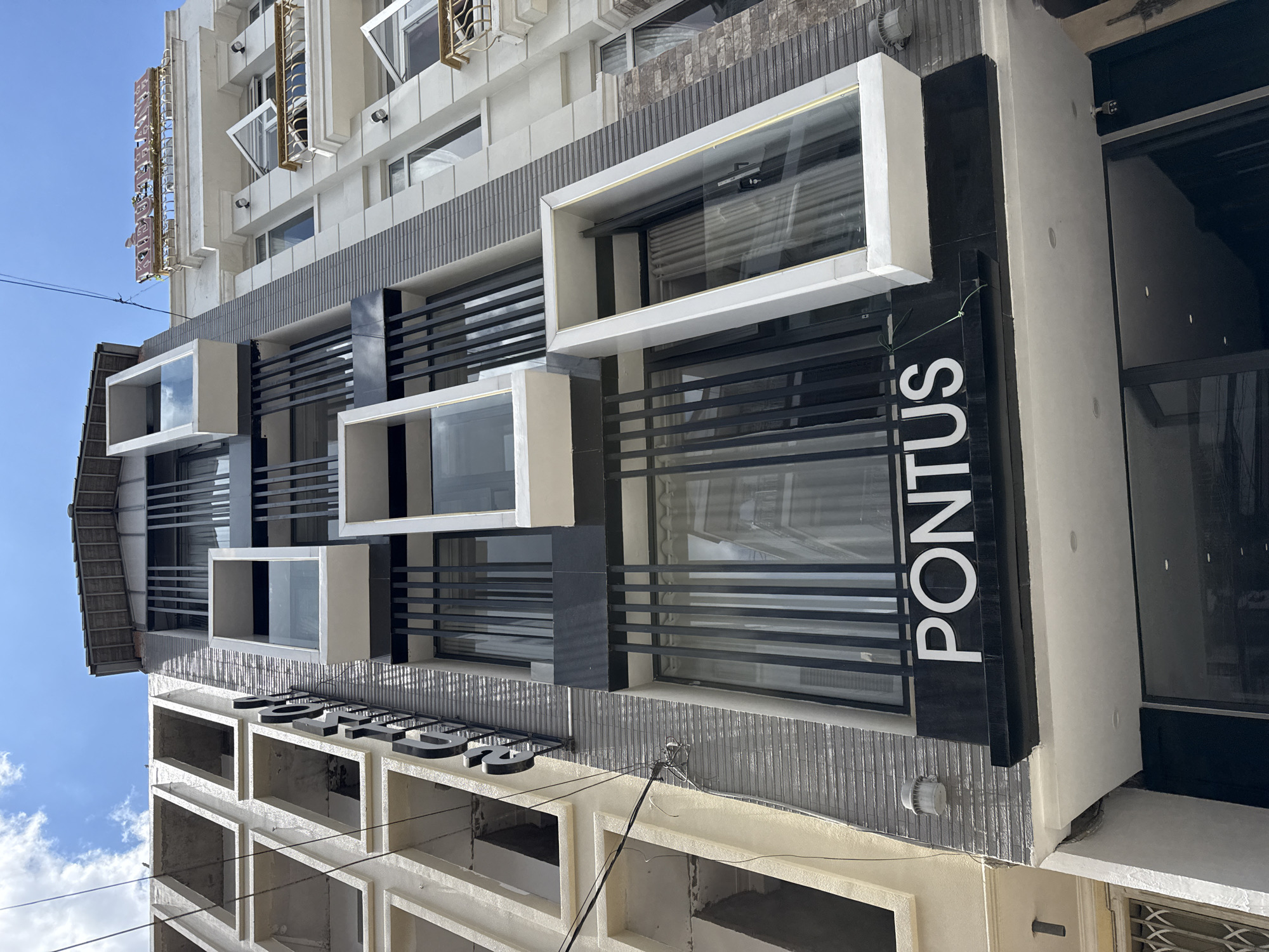 A low-angle, eye-level shot captures the exterior of a modern building complex on a sunny day in Da Lat, Vietnam. The building on the left is partially constructed, with exposed concrete frames for windows. To its right, a gray, textured facade features vertical black slats and several protruding white rectangular window frames with glass. White lettering spelling "PONTUS" is prominently displayed on a black sign below. The building on the far right is cream-colored with white trim and balconies, topped with a red sign that is illegible. The sky above is blue with scattered clouds, and electrical wires are visible.