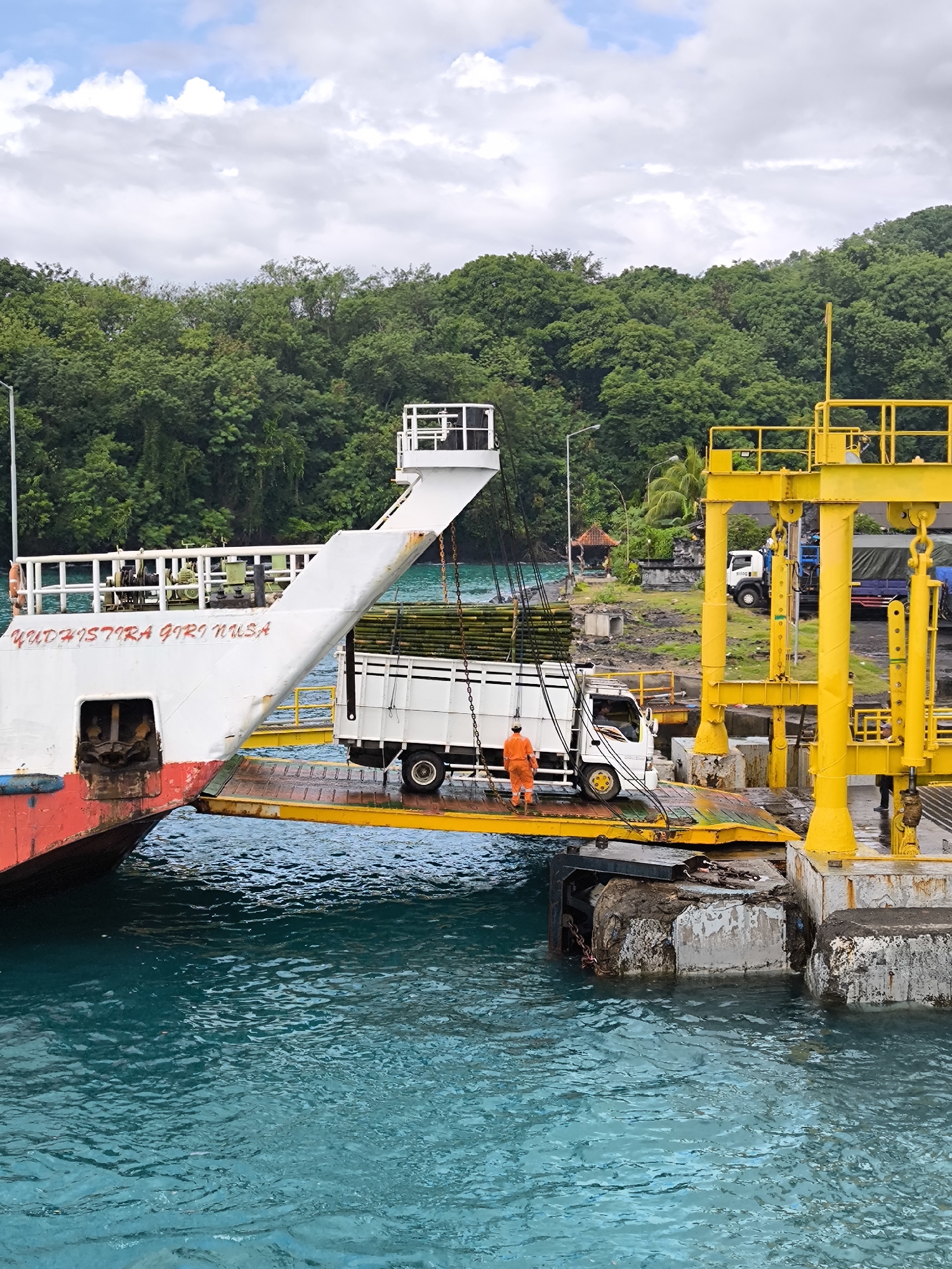 A medium, eye-level shot shows a truck being loaded onto a ferry in an unknown city. The ferry, painted with the name "YUDHISTIRA GIRI NUSA," is docked at a yellow metal loading structure. A white truck carrying what appears to be bamboo or similarly long, green material, is positioned on the ramp connecting the ferry and the dock. An individual wearing an orange jumpsuit stands near the truck's rear wheels. In the background, a dense forest is visible, along with glimpses of buildings and parked vehicles on the dock. The water surrounding the dock is turquoise. The sky is overcast.