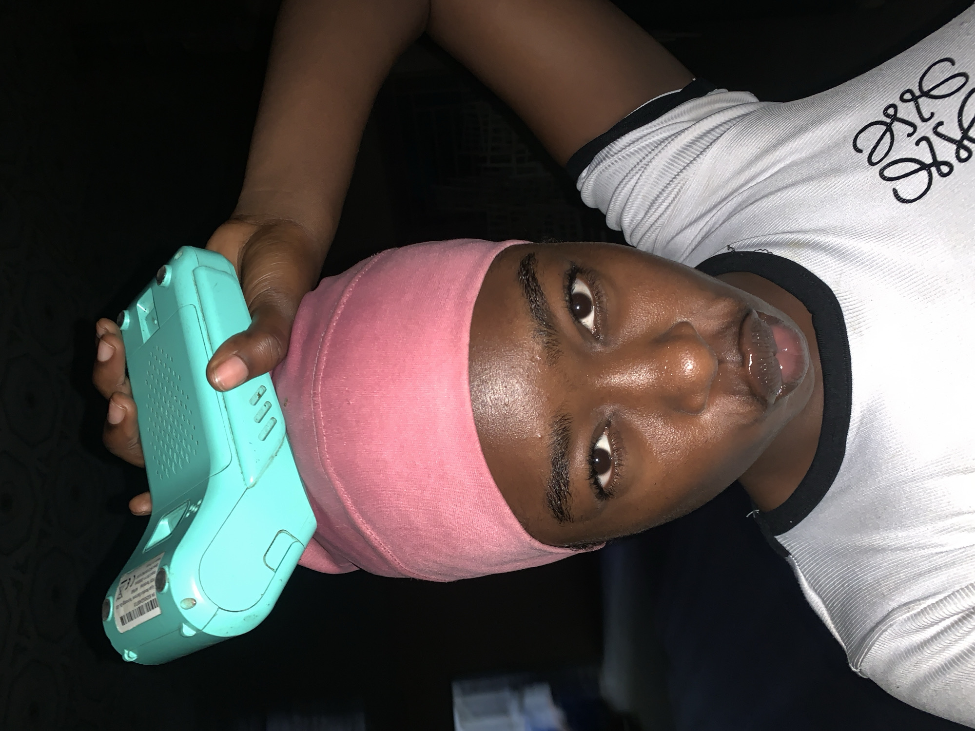 A person with dark skin wearing a pink head covering and a white shirt with black trim is holding a blue handheld device in their left hand. The person is looking directly at the camera with their mouth slightly open. The background is dark and indistinct. The specific location is not discernible from the image alone, and no elements indicate it is in New Stadium, Nigeria.