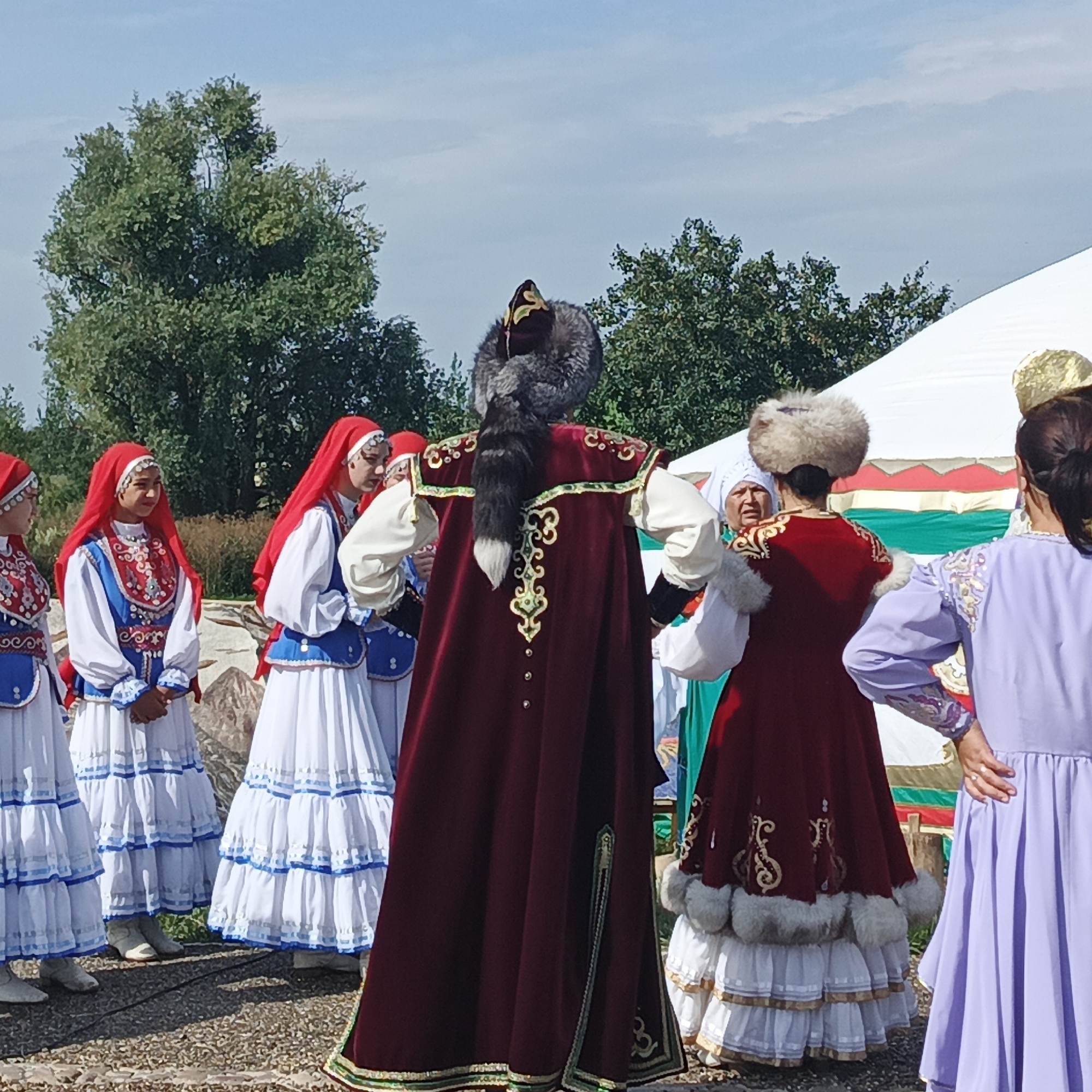 This is a full shot of a group of people in traditional attire standing outdoors. The setting appears to be a public event, possibly a festival, given the presence of a large white tent with colorful fabric panels behind the individuals. A few trees with green foliage are visible in the background, and the sky is blue with scattered clouds.
In the foreground and center, a person wearing a deep red velvet coat with ornate gold embroidery and a fur-trimmed collar is facing away from the camera. This coat is worn over a white shirt with wide sleeves and a white tiered skirt with gold trim. A large fur hat with a prominent tail hangs down the back of the coat.
To the left of this central figure, three women are standing, all wearing white dresses with blue vests and red headscarves. The dresses have tiered skirts with blue and white trim. Their vests are decorated with intricate embroidery in red, blue, and white. The woman on the far left is mostly out of frame, while the other two are clearly visible, looking towards the central figure.
To the right of the central figure, another person in a similar red velvet coat with fur trim is standing, also with their back mostly to the camera. This individual is wearing a fur hat. Next to them, a woman in a light purple dress is seen from the side, her hair pulled back in a ponytail. She has on a gold headdress. Another person, partially visible to the left of the purple-dressed woman, appears to be wearing a white headdress.
The ground in front of the people is a textured surface, possibly asphalt or gravel, with some shadows cast by the figures and the tent. The overall impression is a cultural gathering, likely in a rural or semi-rural area of Russia, possibly in Nikolo-Beryozovka, given the context provided.